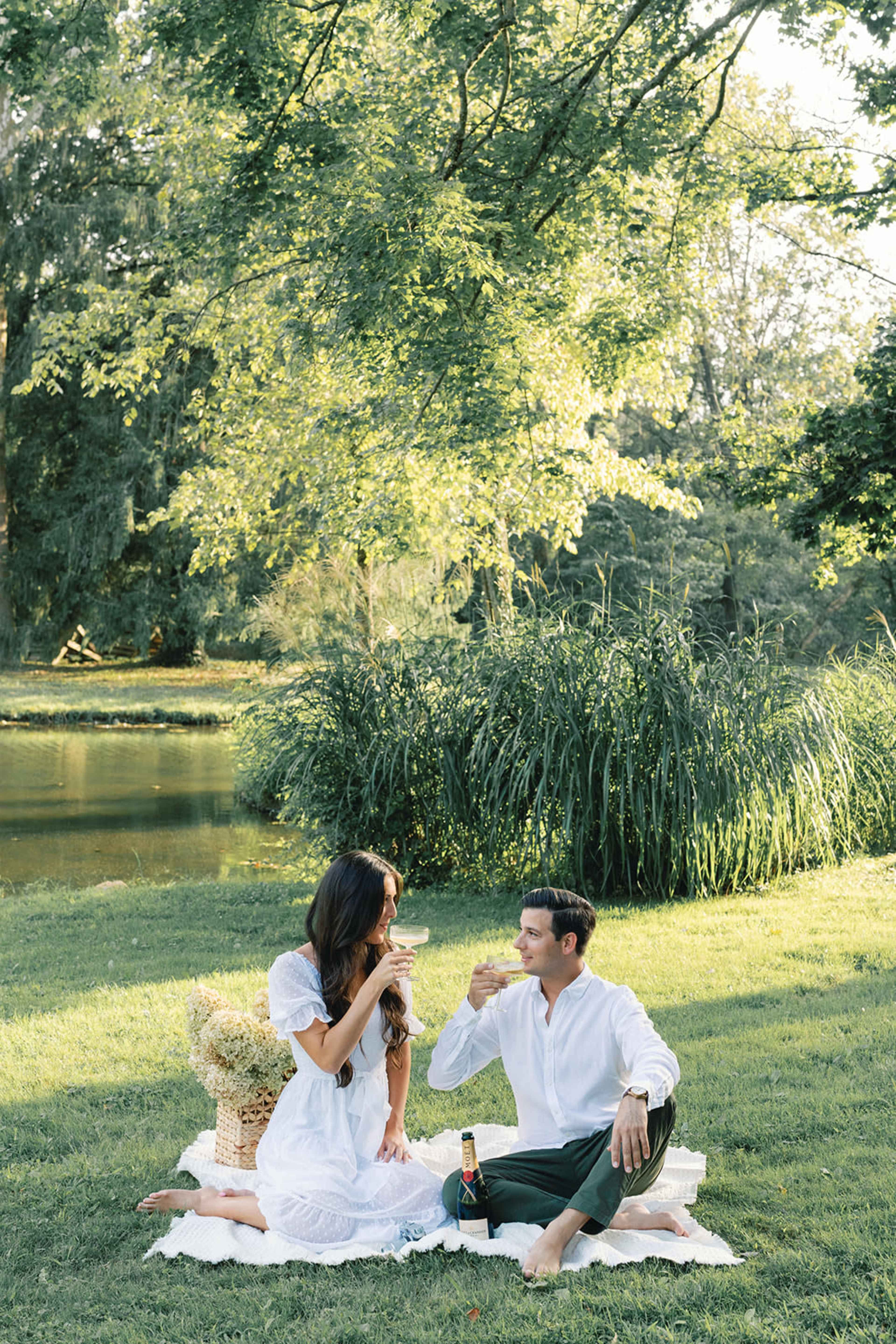A couple sits on a blanket by a pond, enjoying drinks and surrounded by greenery.