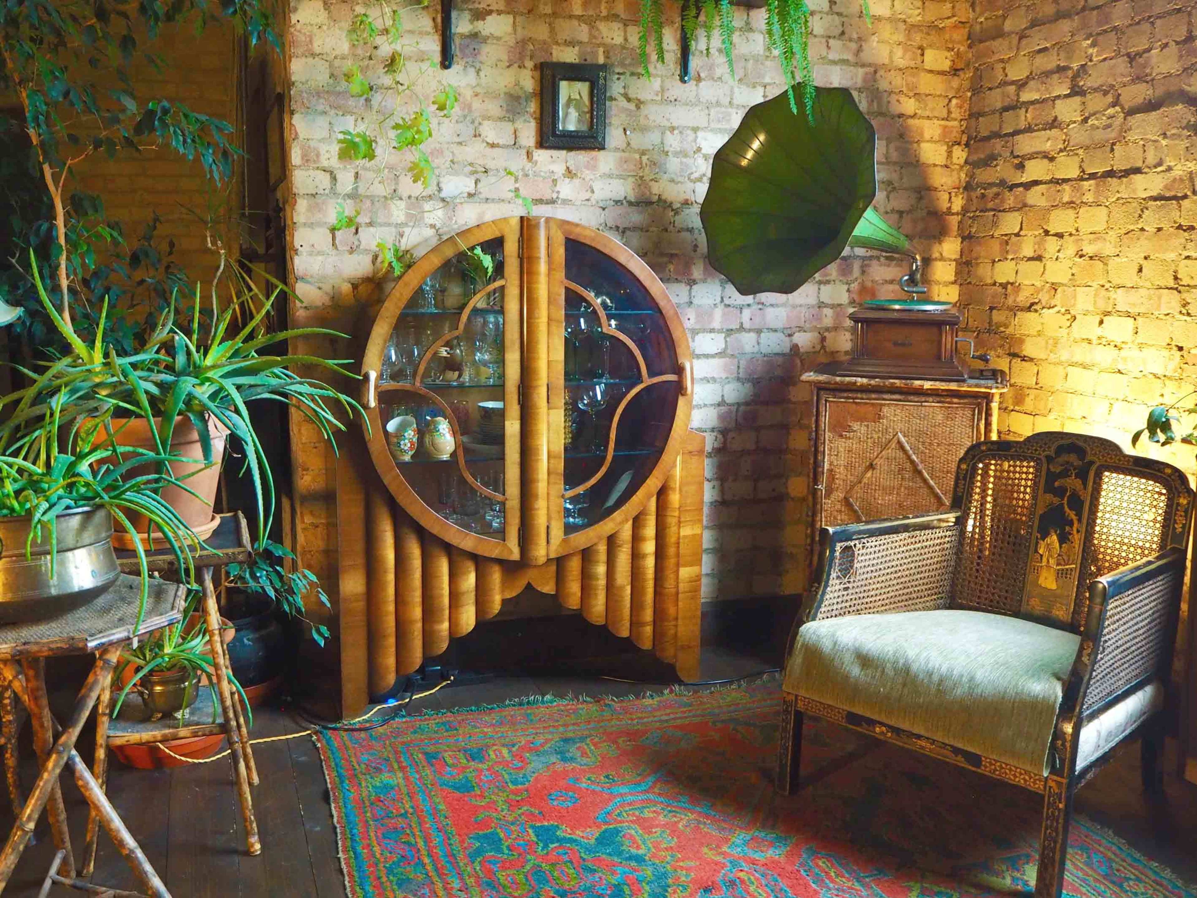 A cozy corner of a room featuring a wooden cabinet with glass displays, an ornate chair, and various plants against a textured brick wall.