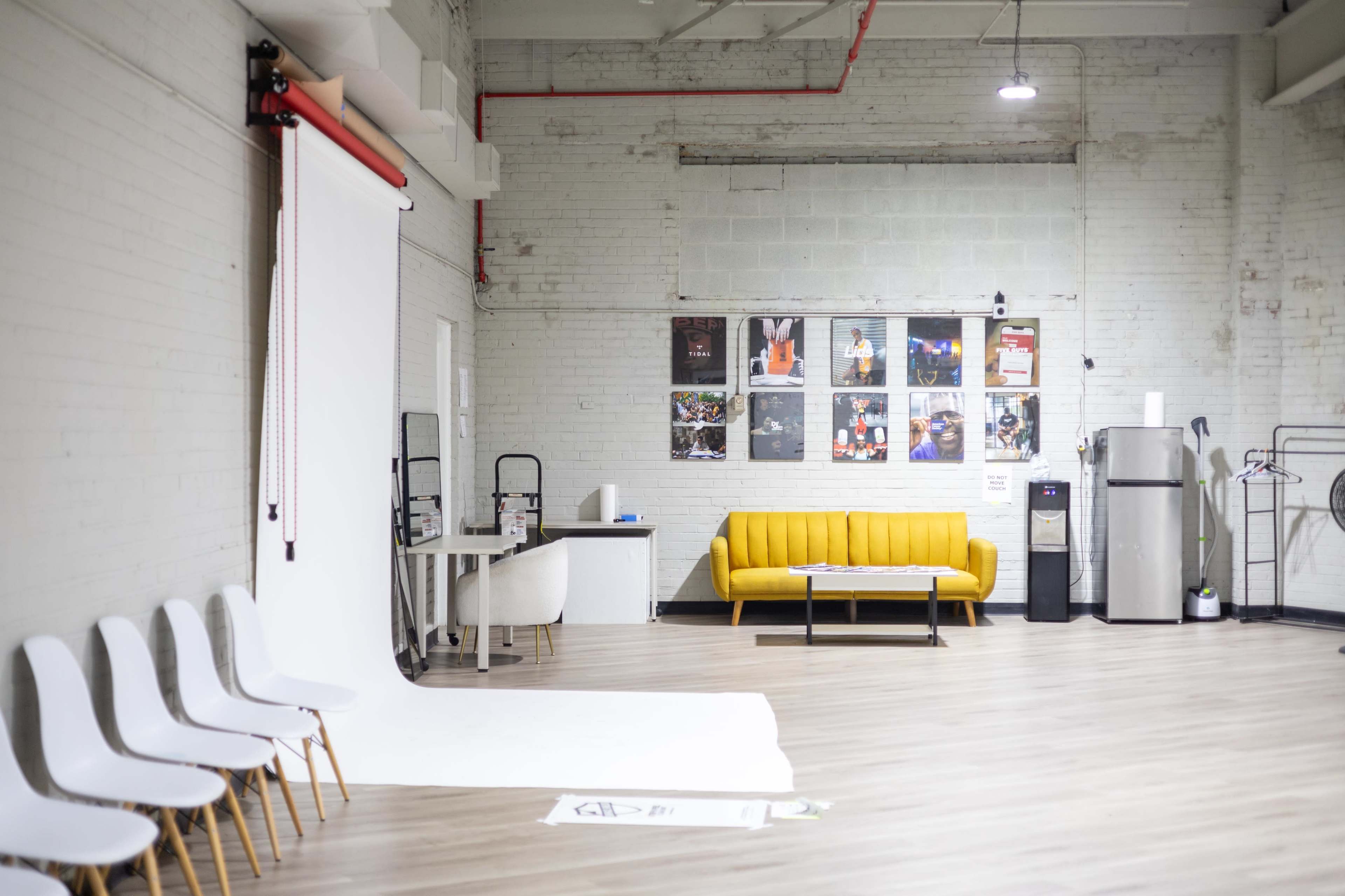 The image shows a spacious studio with a yellow sofa, a large roll of white backdrop paper, and several posters on the wall.