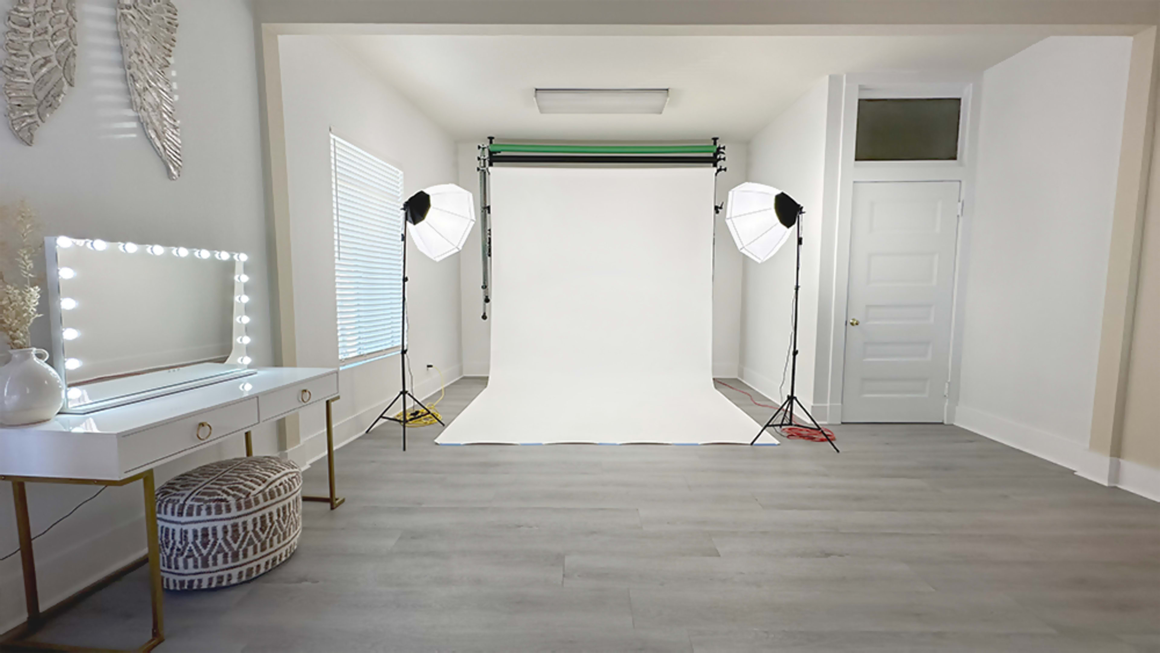 A well-lit photography studio features a white backdrop, two softbox lights, and a mirror vanity in a minimalistic room.