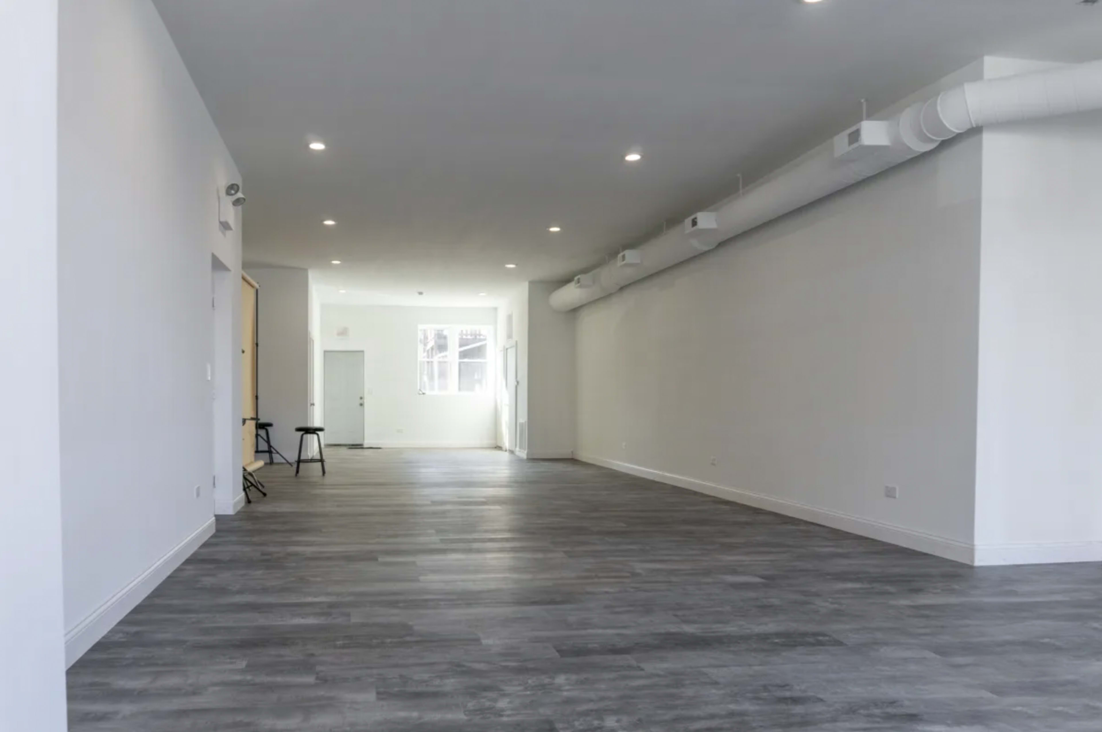 A spacious, empty room with grey flooring and white walls, featuring overhead lighting and windows at one end.