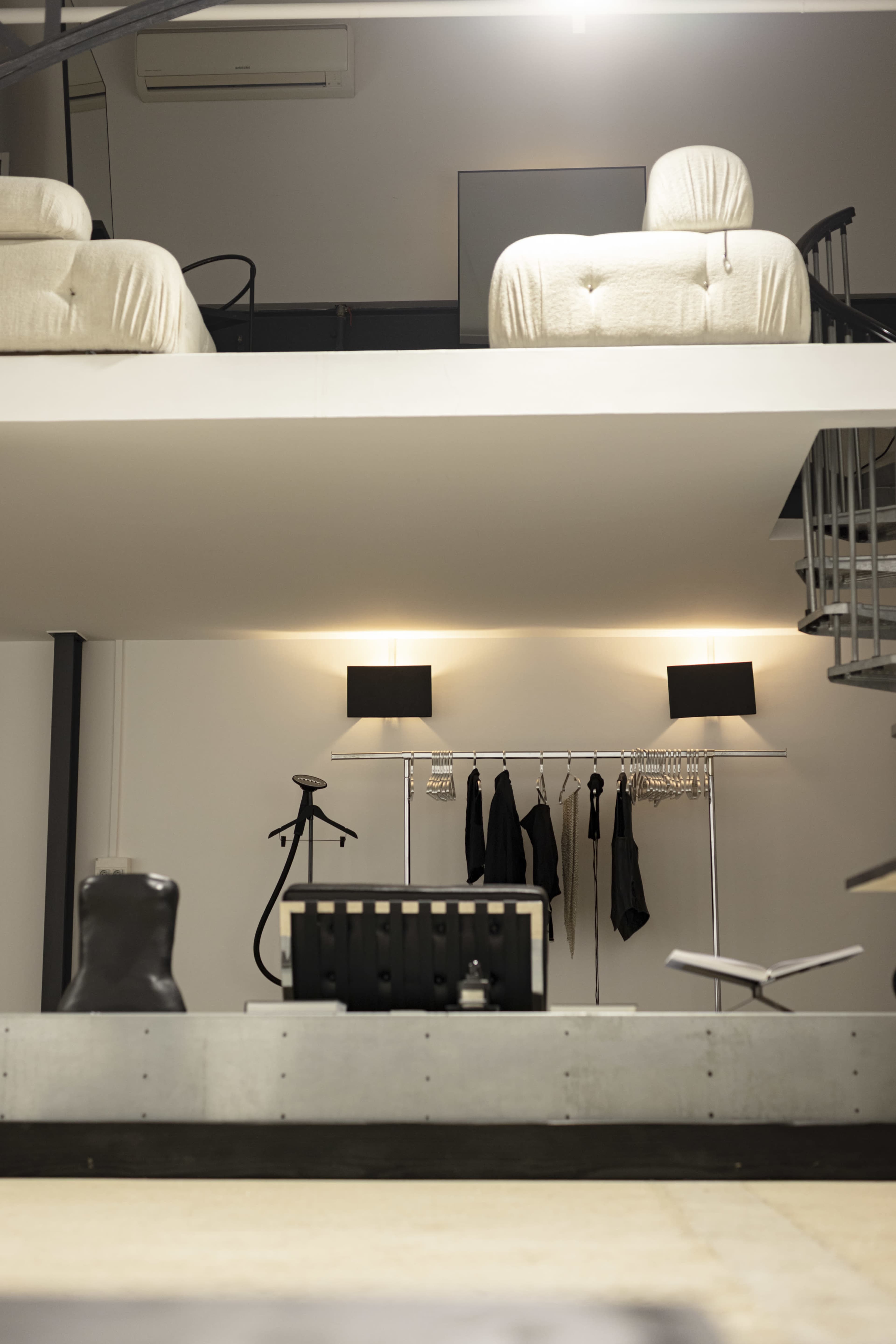 The image features a minimalist interior with two upholstered chairs on a balcony above, a clothing rack with black garments, and a staircase leading to the upper level.