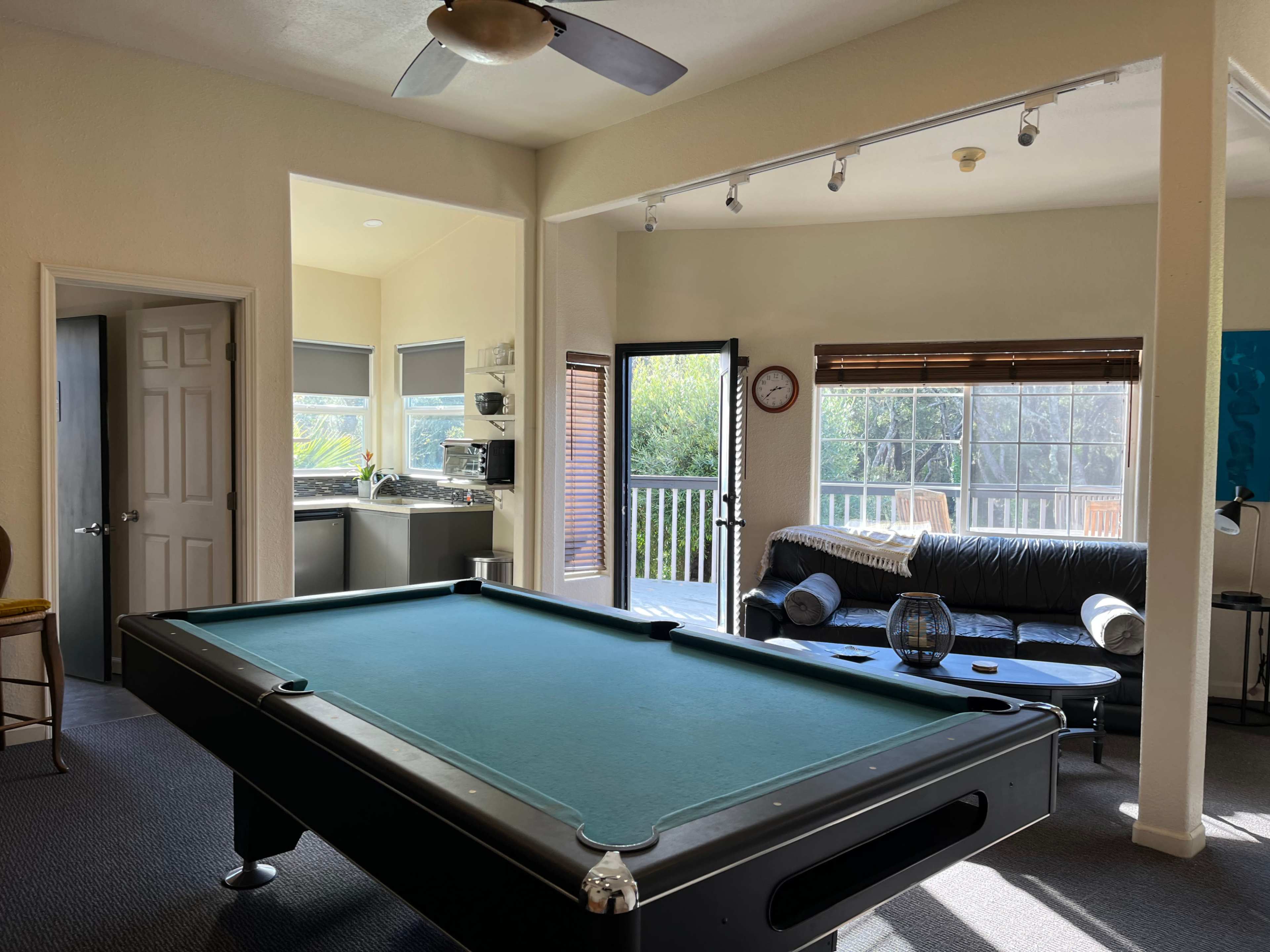The image shows a spacious living area with a pool table, a sofa, and a kitchen accessible through an open doorway.