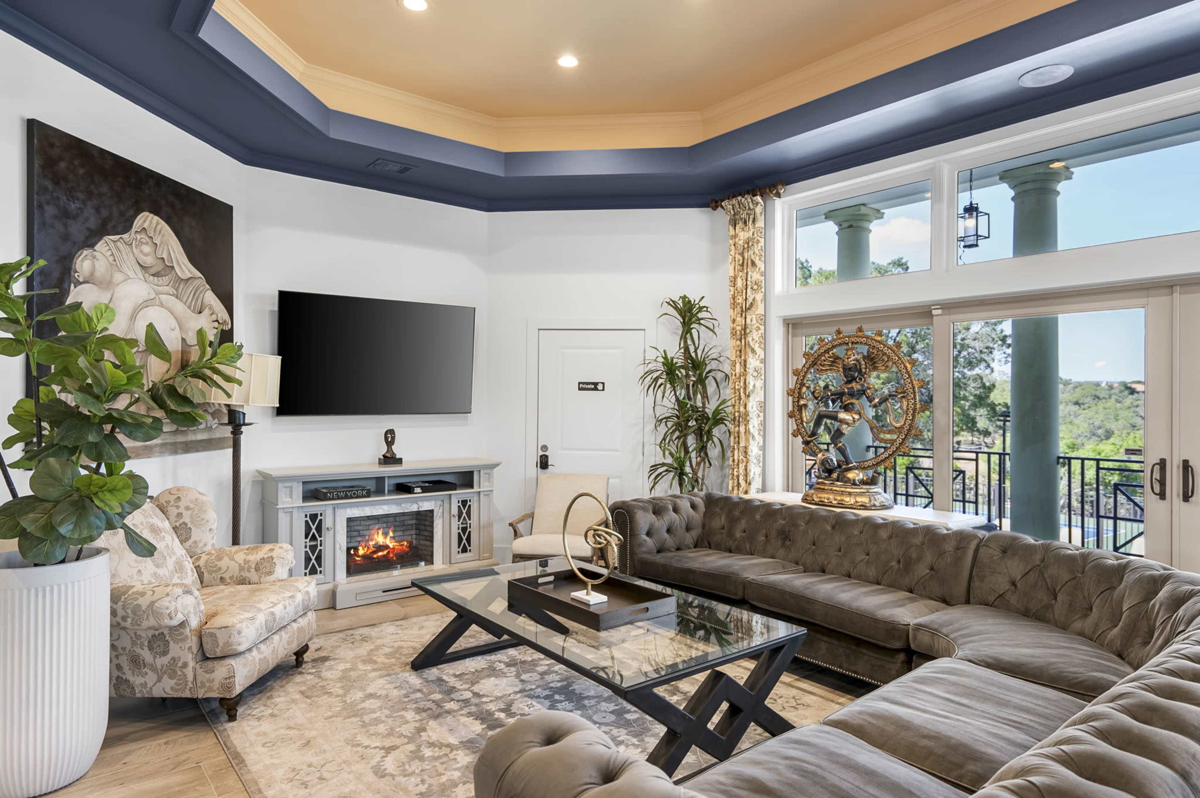 The image shows a modern living room featuring a large tufted sectional sofa, a glass coffee table, an entertainment center with a television, and decorative plants, all set against a backdrop of large windows.