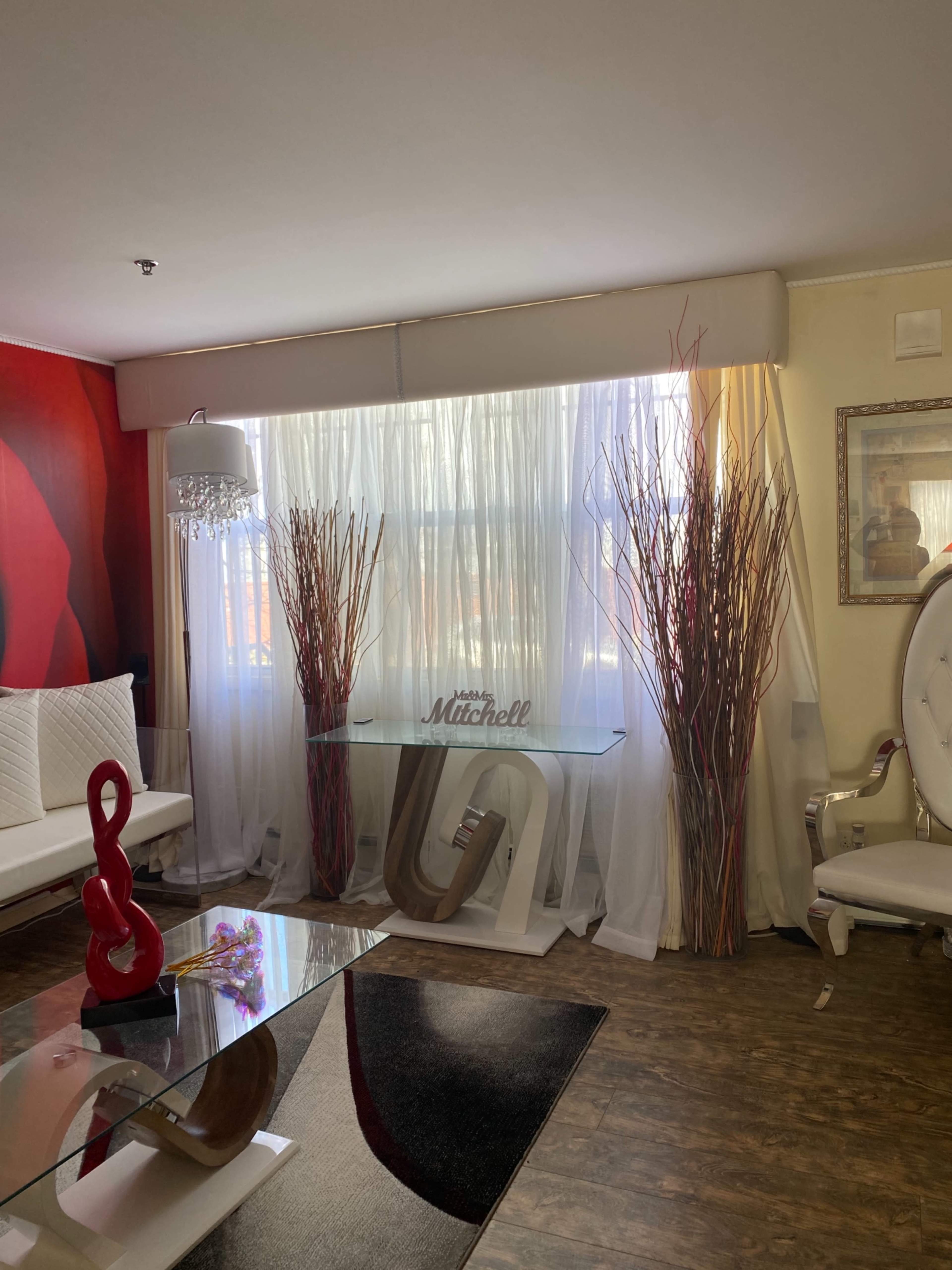 A living room with a white sofa, a glass coffee table, tall decorative branches in vases, and a console table in front of large sheer curtains.