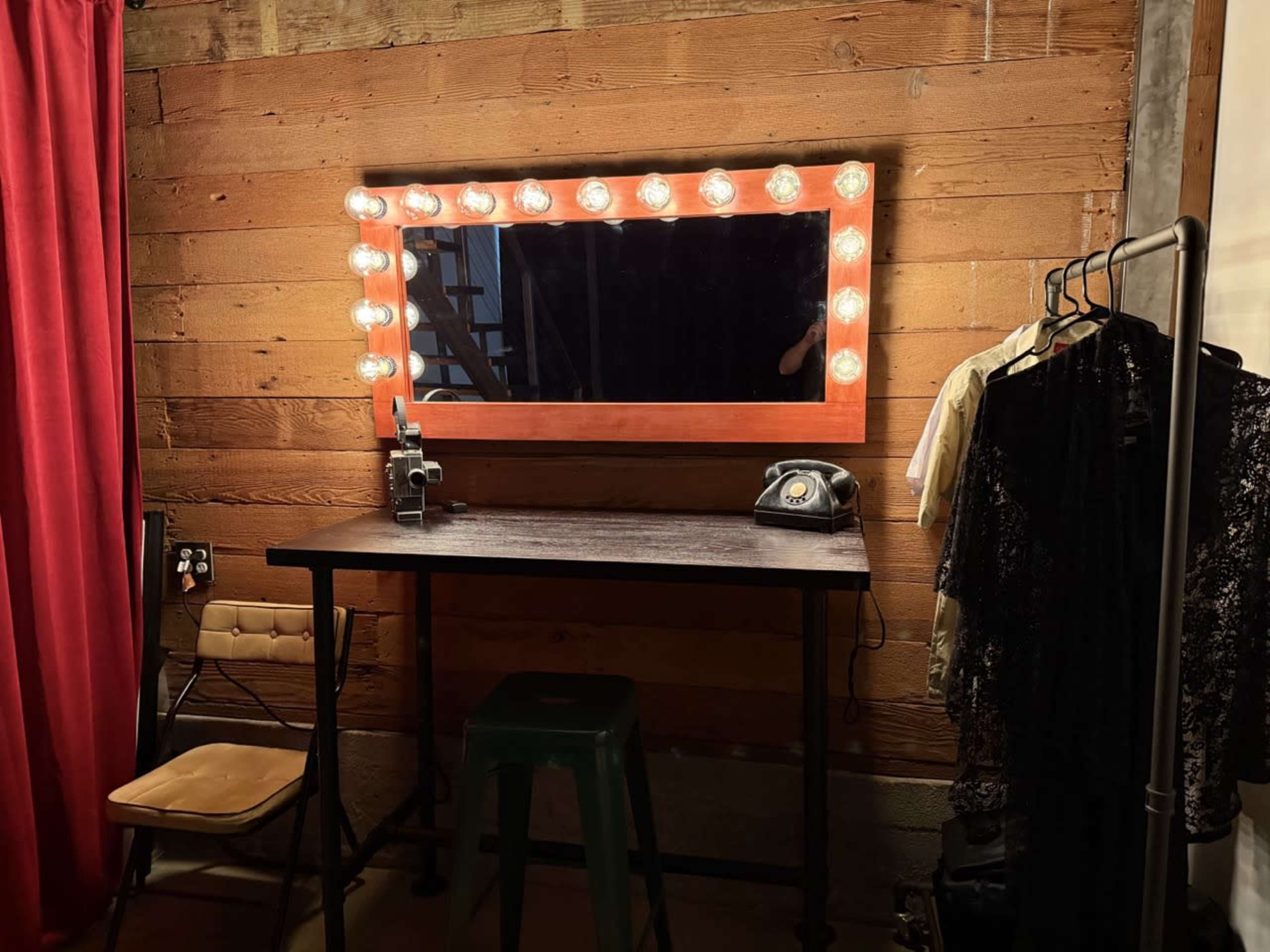 The image shows a stylist's vanity area with a mirror framed by lights, a vintage telephone, and clothing hanging on a rack against a wooden wall.