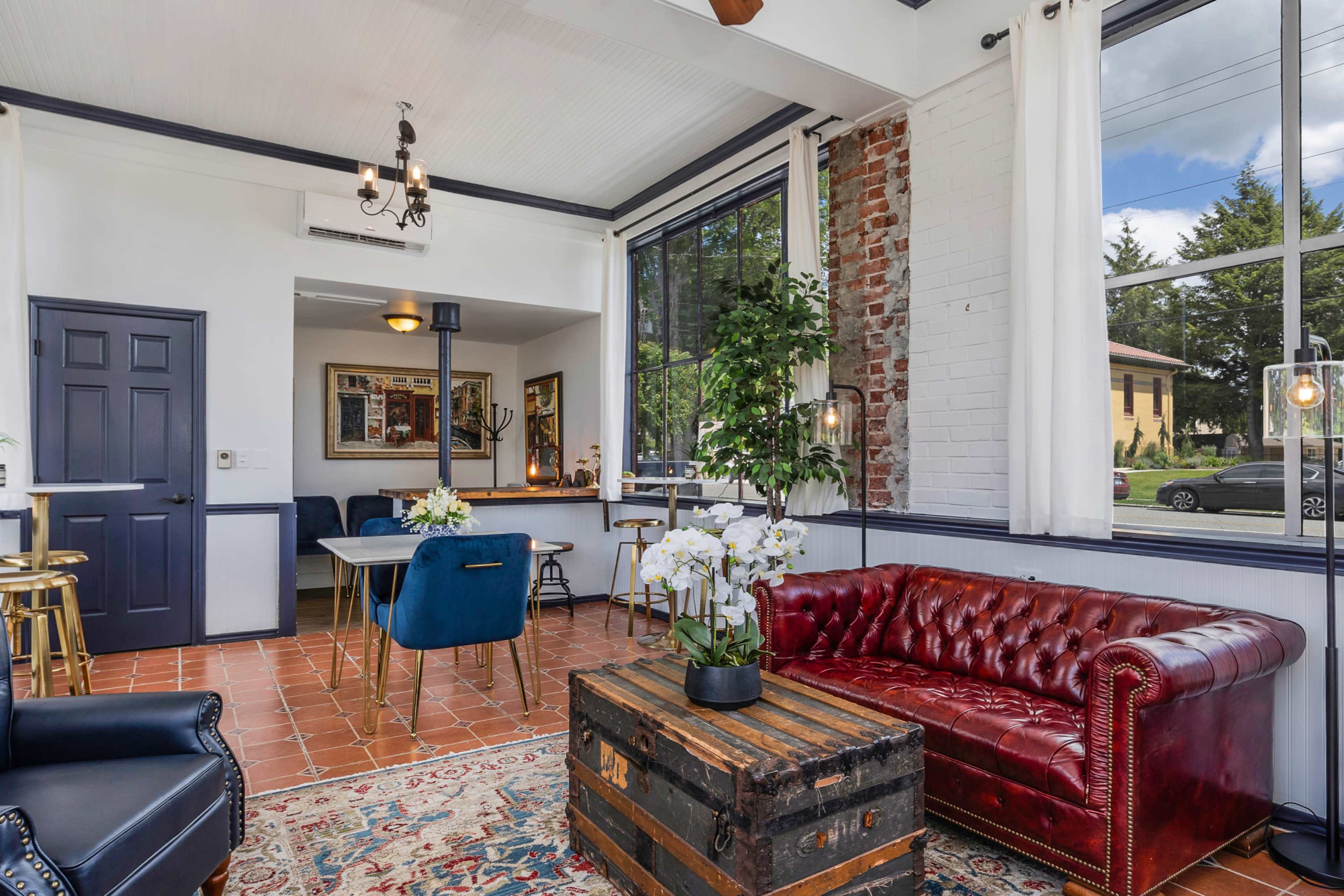 A cozy lounge area features a red leather sofa, a wooden coffee table, and a mix of seating options with large windows allowing natural light to fill the space.