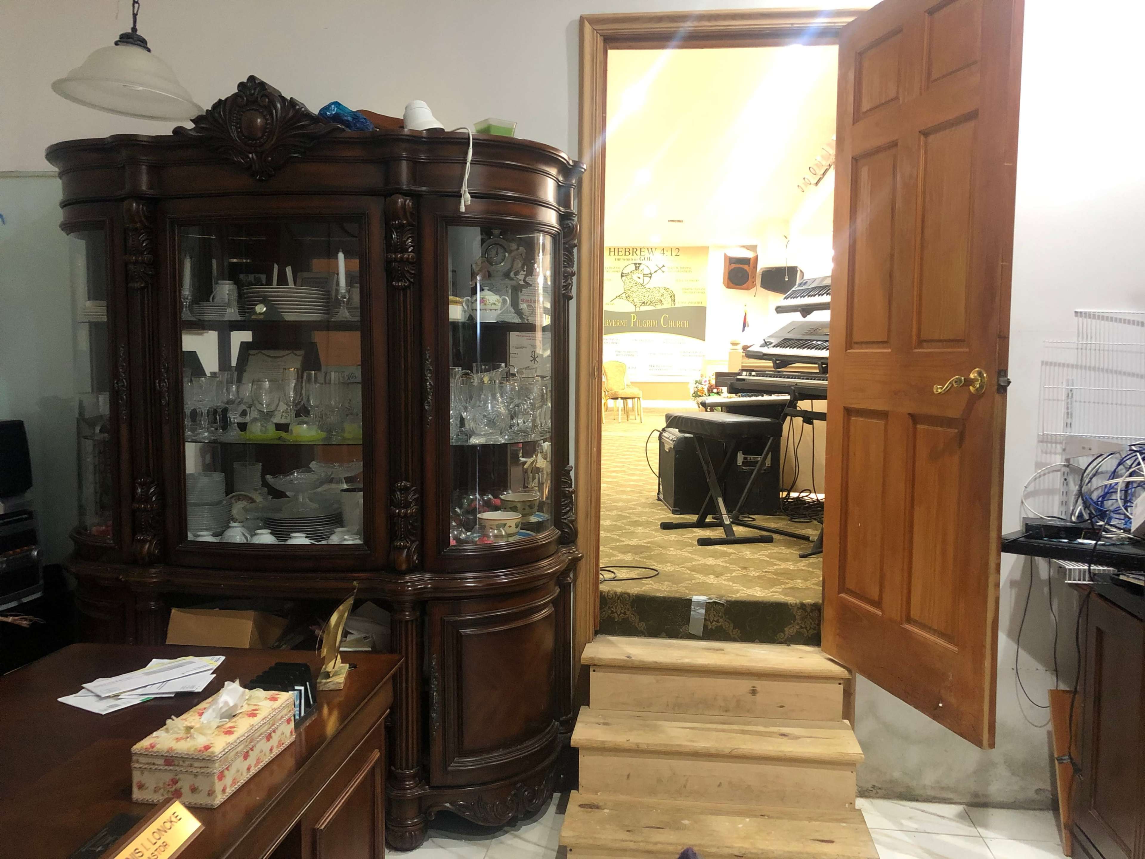 A wooden cabinet with glass doors displaying glassware, next to an open door leading to a room with musical instruments and carpeted steps.