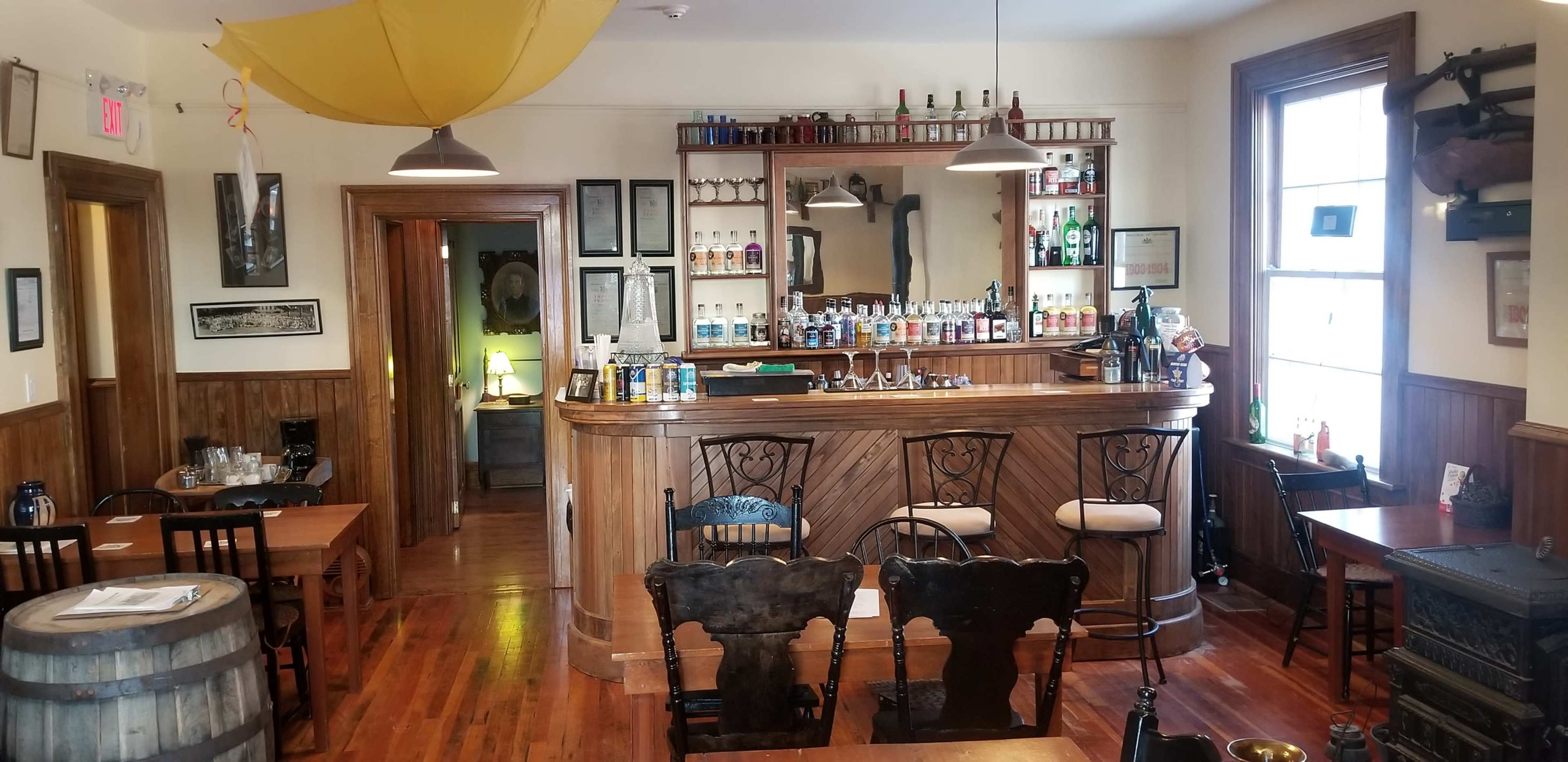 A well-furnished bar area with a wooden counter, several rows of bottles on shelves, and wooden tables and chairs arranged around the space.