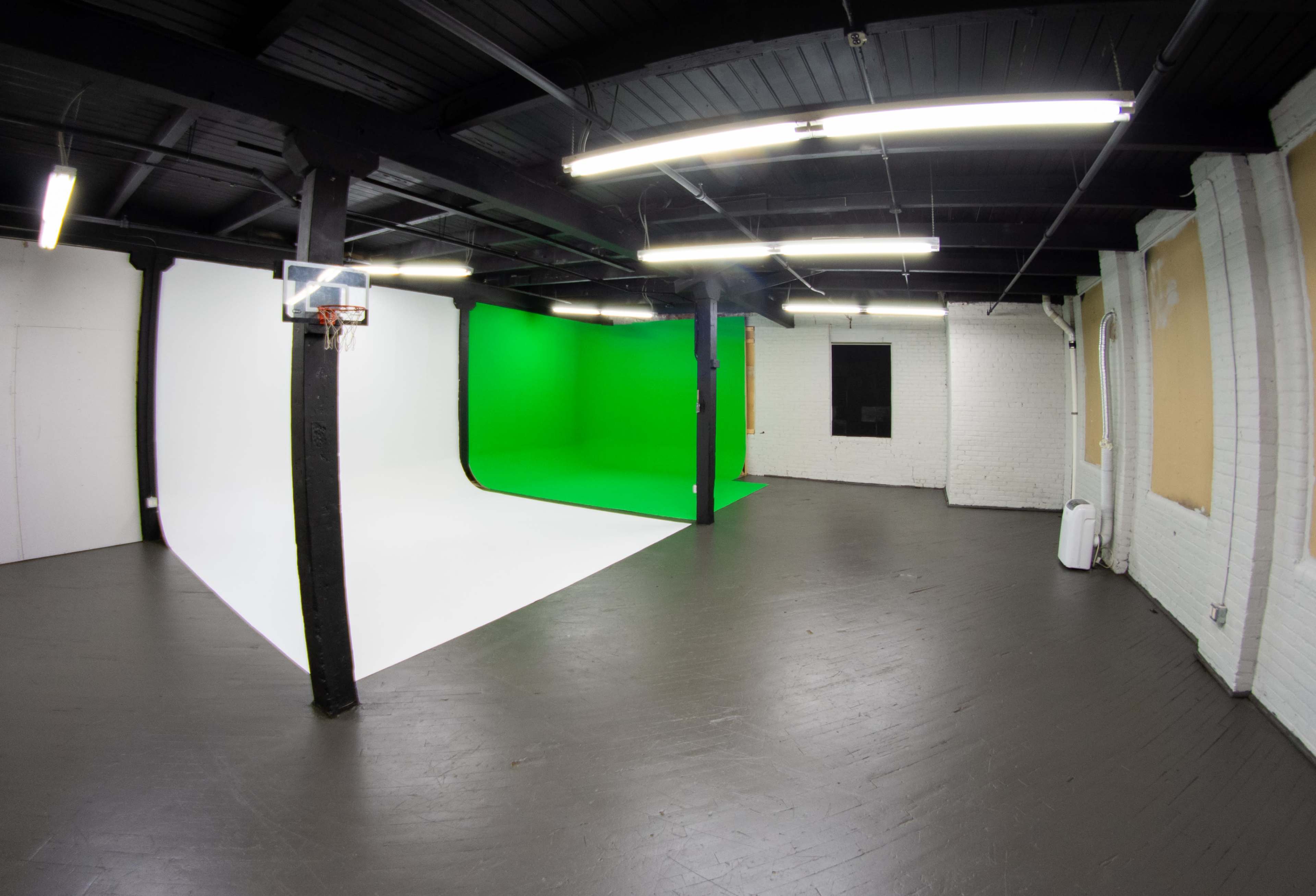 A spacious studio with a white wall and a green screen backdrop, featuring black beams and fluorescent lighting.