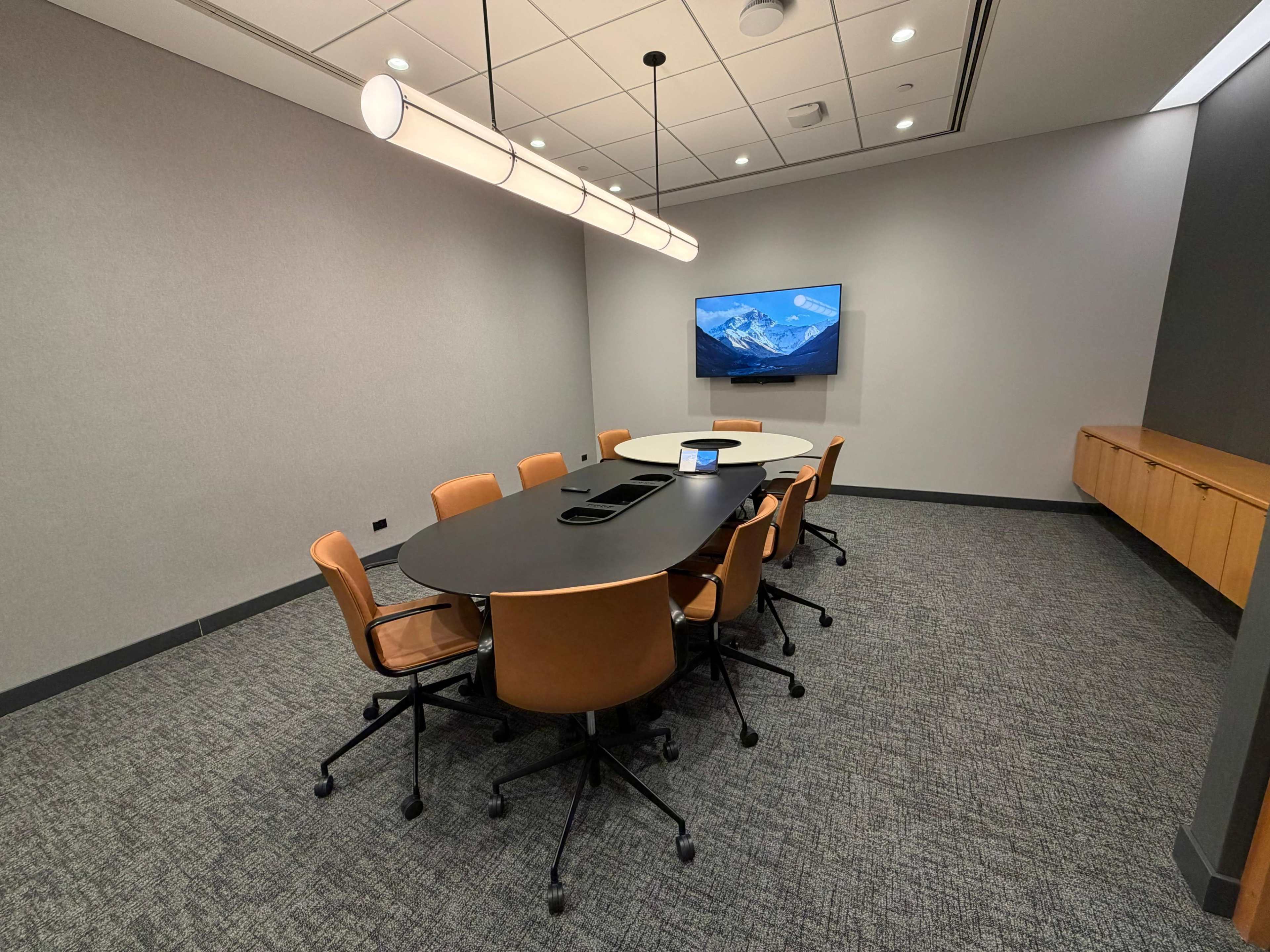 A modern conference room features a large oval table surrounded by brown chairs and a wall-mounted screen displaying a mountain landscape.
