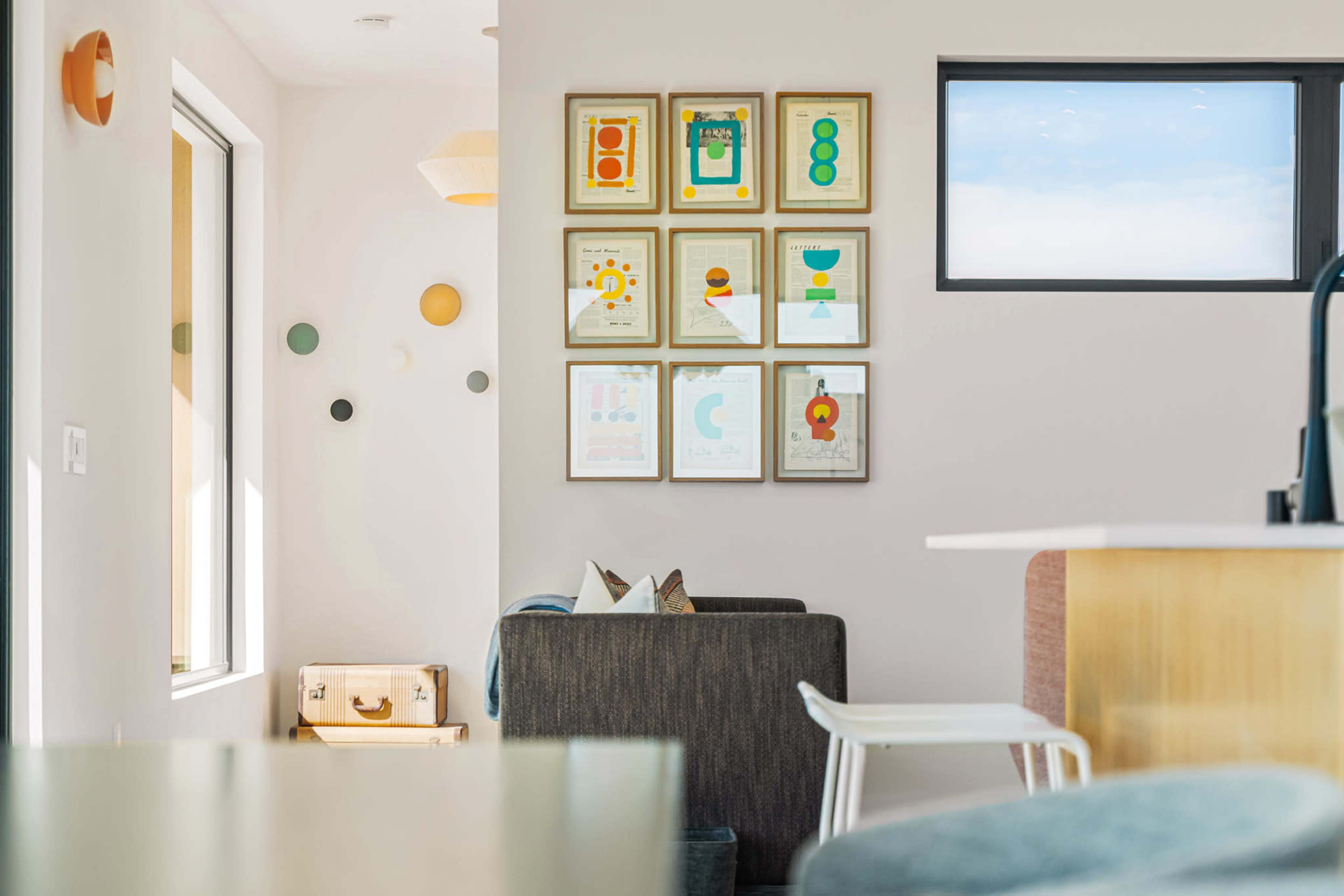 The image shows a modern interior space featuring a framed gallery of colorful artwork on a light-colored wall, a gray couch, and a view of large windows with natural light.