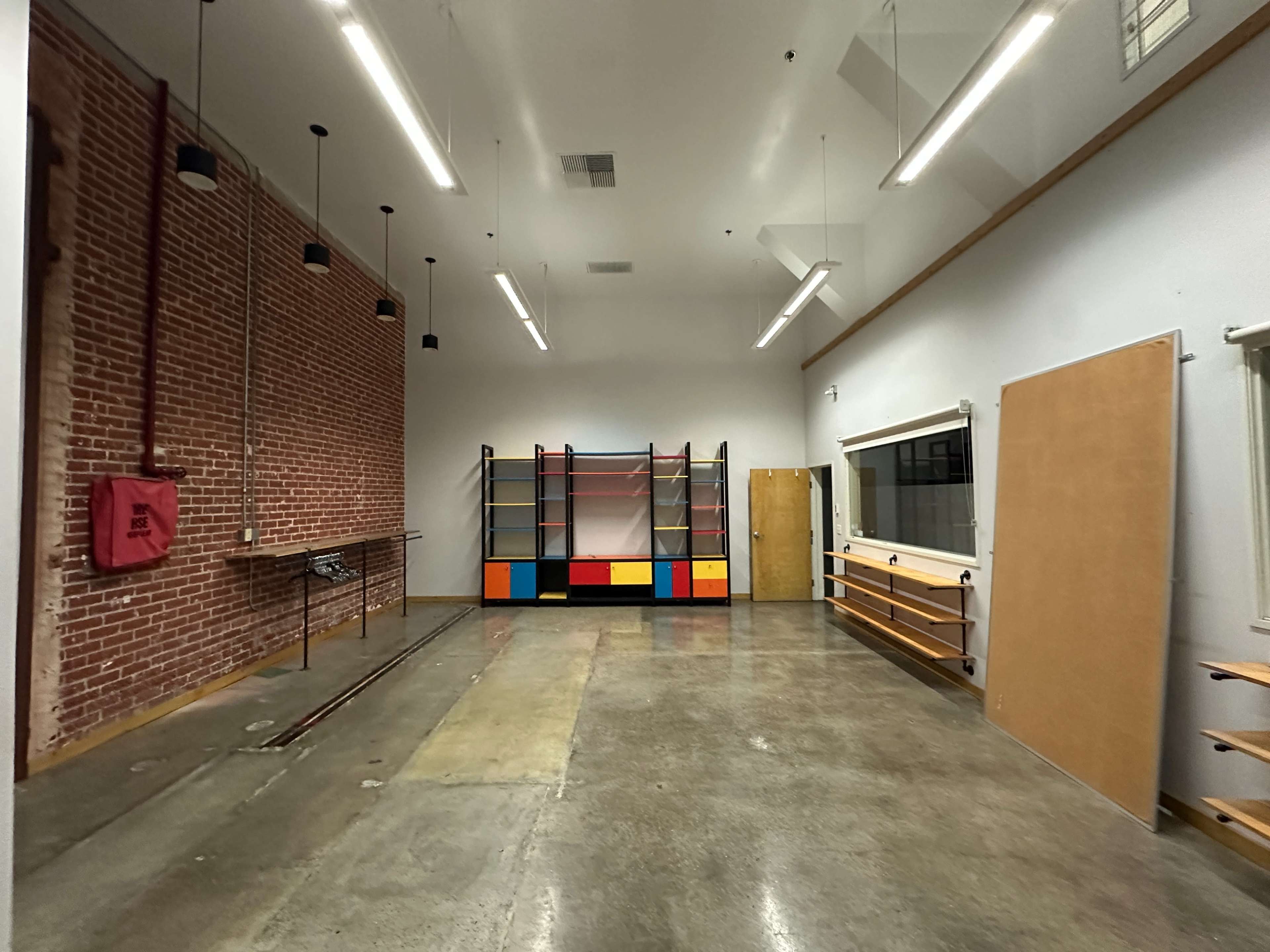 The image shows an empty room with a brick wall on one side, a colorful shelving unit at the center, and various wooden shelves mounted on the opposite wall.