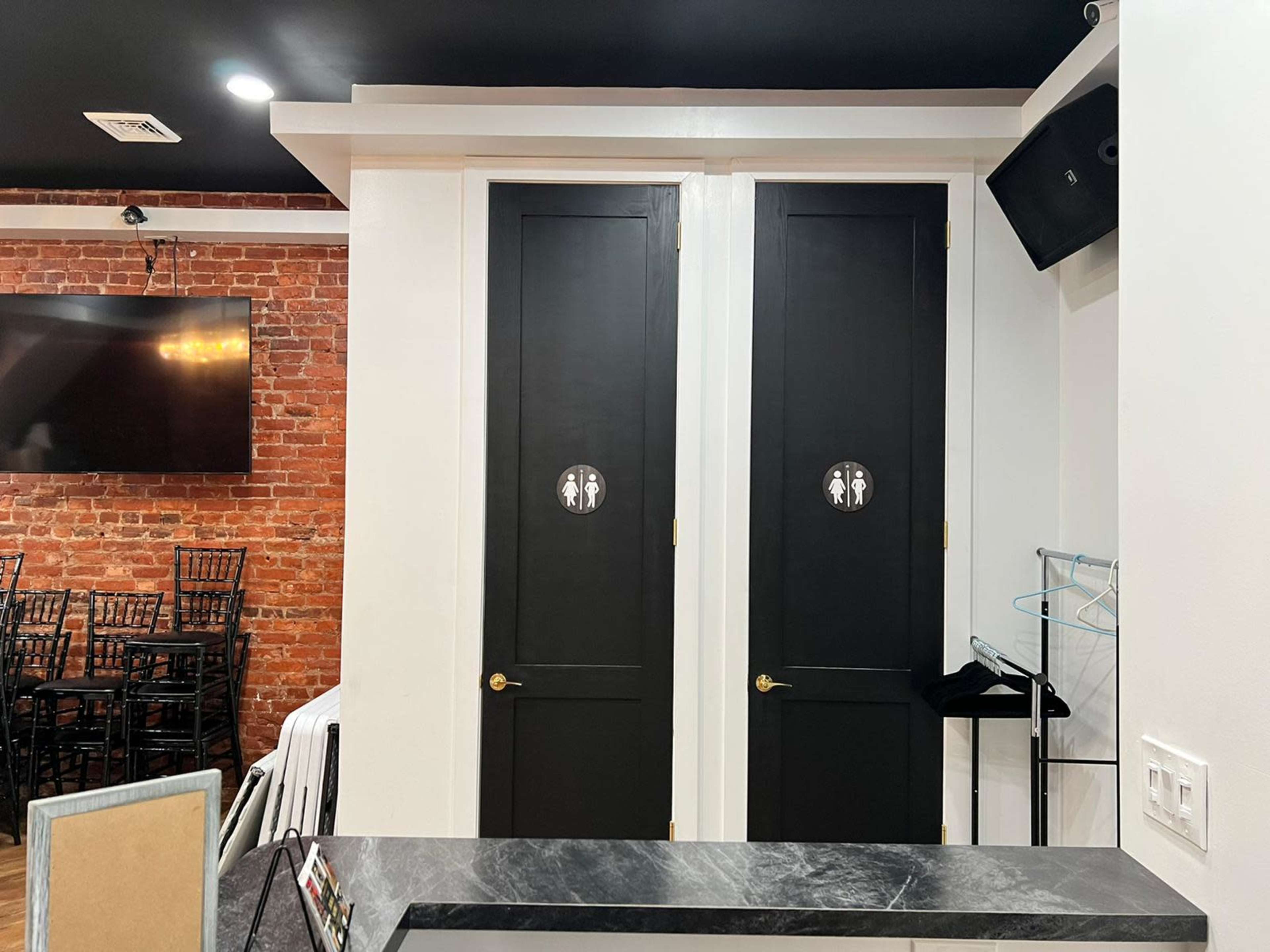 Two black doors with circular signs indicating a restroom, set against a backdrop of exposed brick and a television.