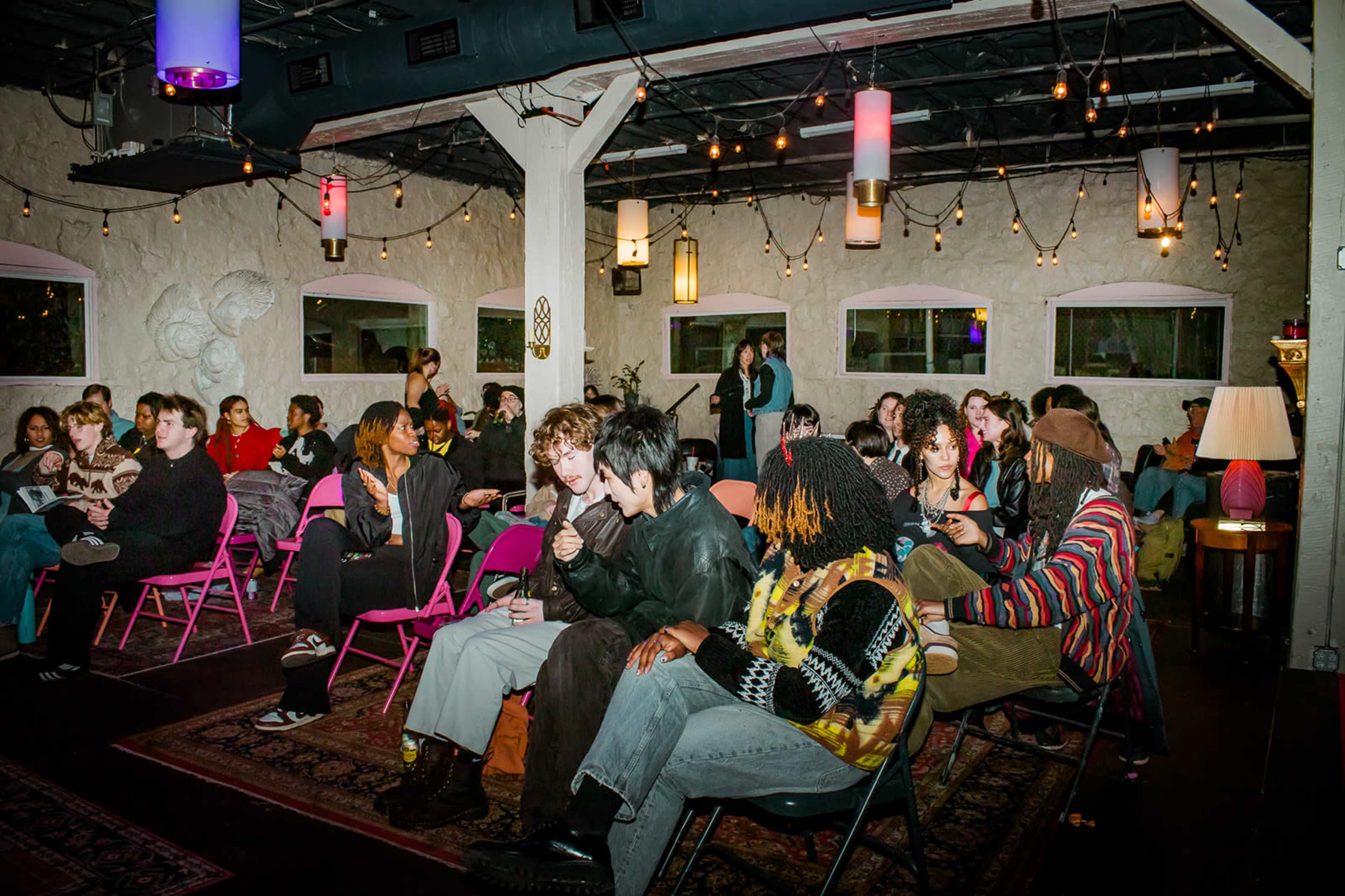 A diverse group of people is seated in a softly lit room adorned with string lights and colorful lanterns, engaged in conversation during an event.