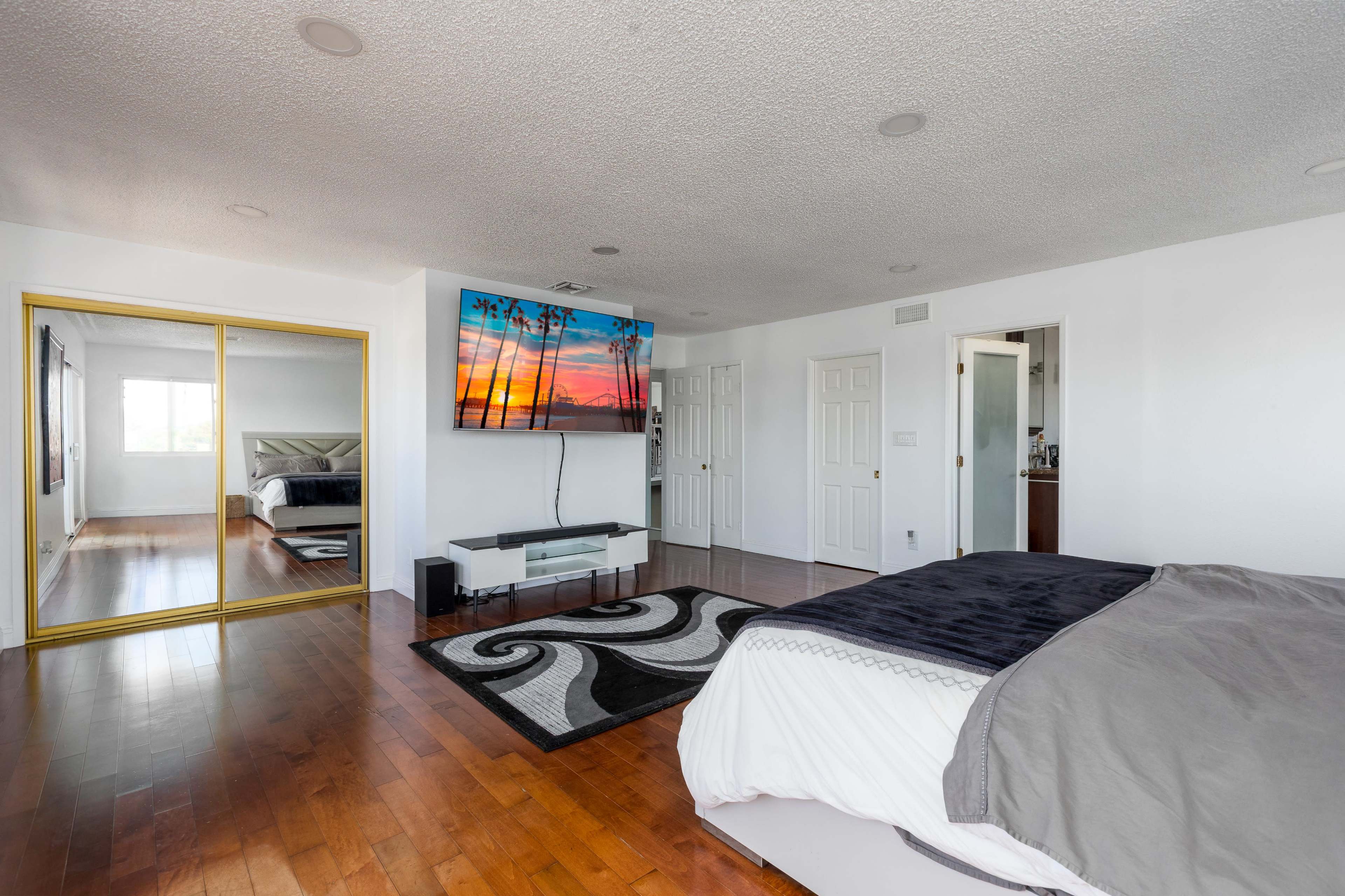 The image shows a spacious bedroom with wooden flooring, a large mirrored closet, a flat-screen TV mounted on the wall, and a bed with gray bedding.