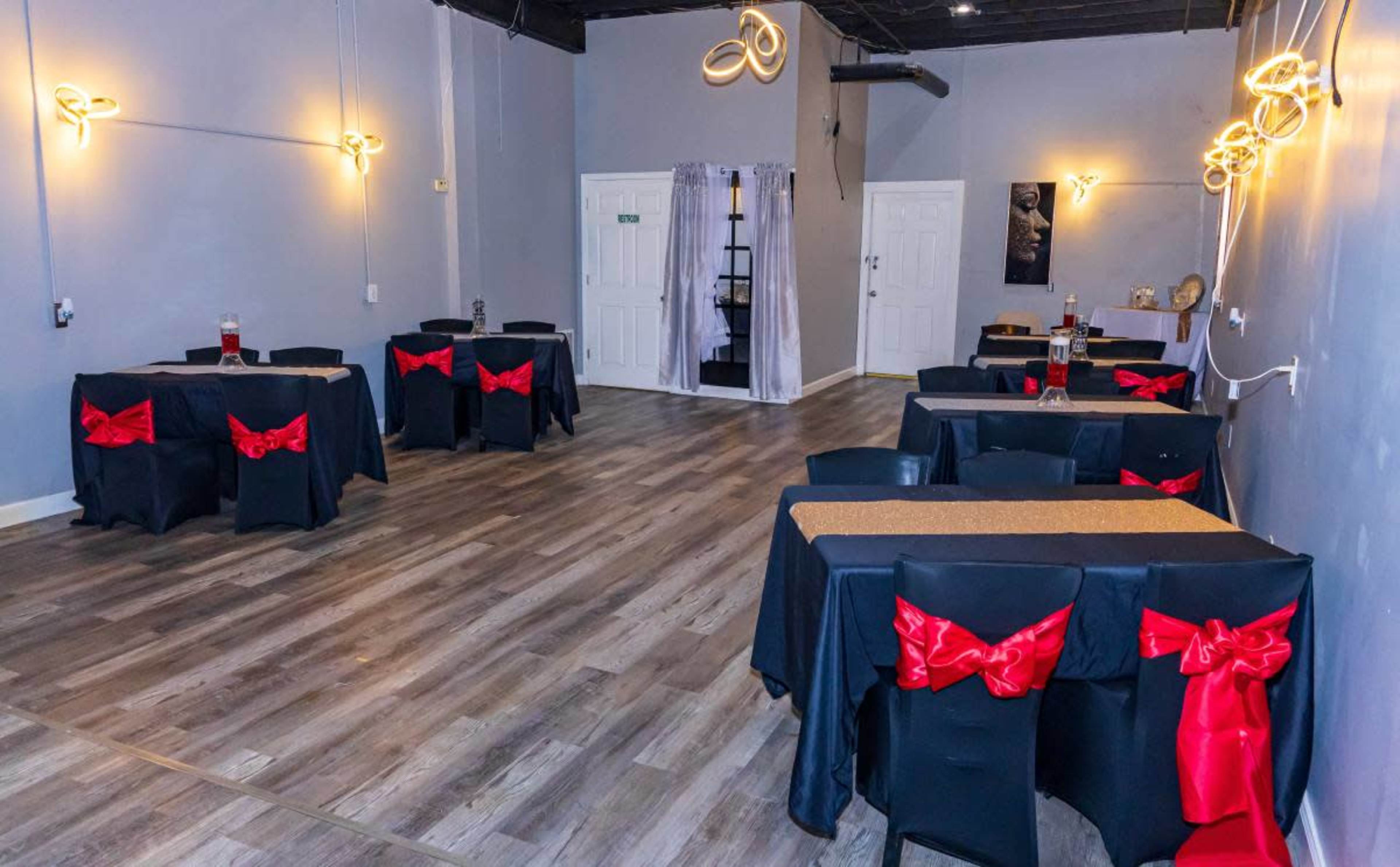 The image shows a spacious, empty event venue with black tablecloths adorned with red bows, wooden flooring, and decorative lighting.