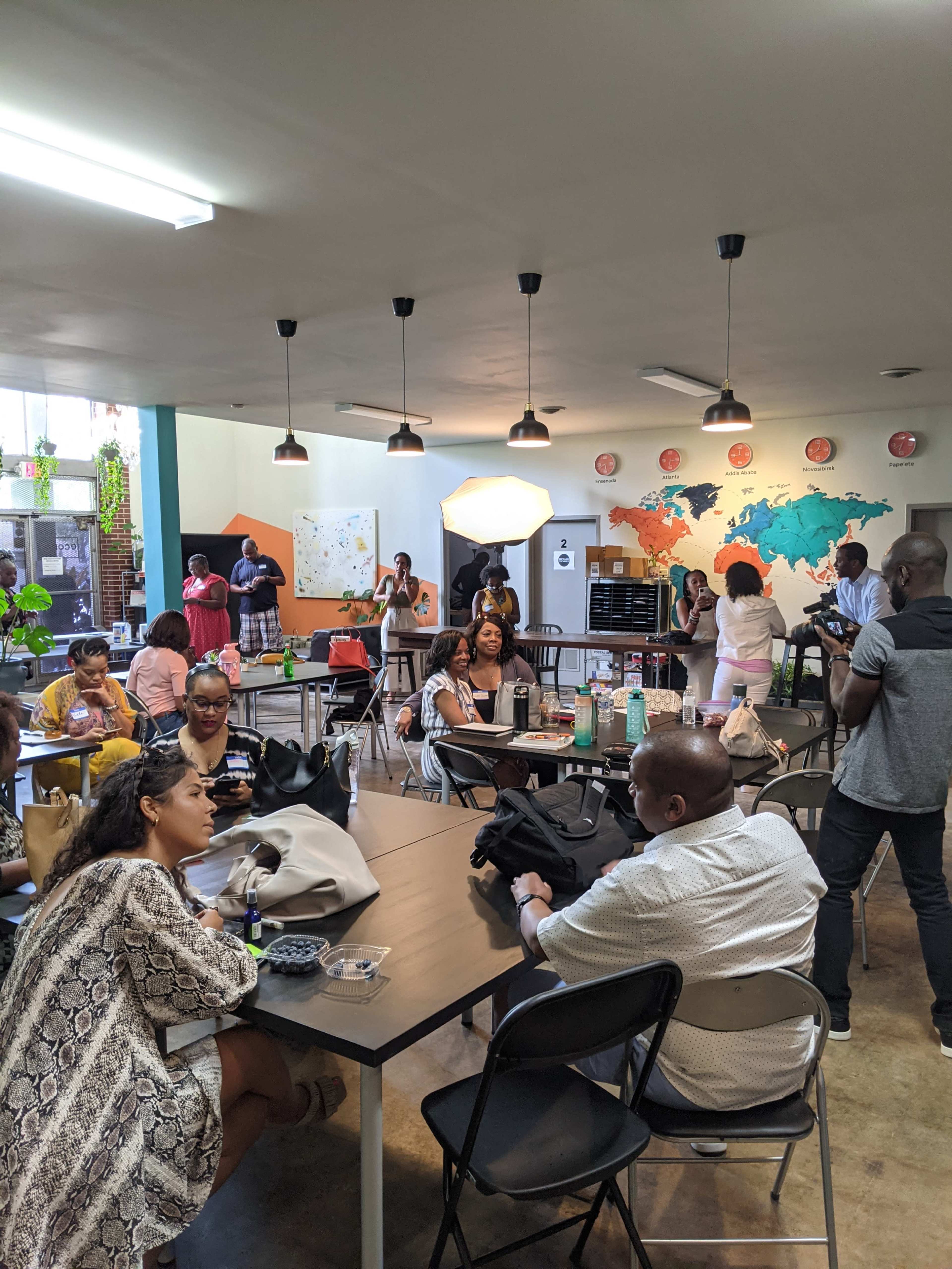 A diverse group of people are seated and standing in a large, brightly lit room with tables, discussing and engaging with each other amidst a backdrop of a world map.