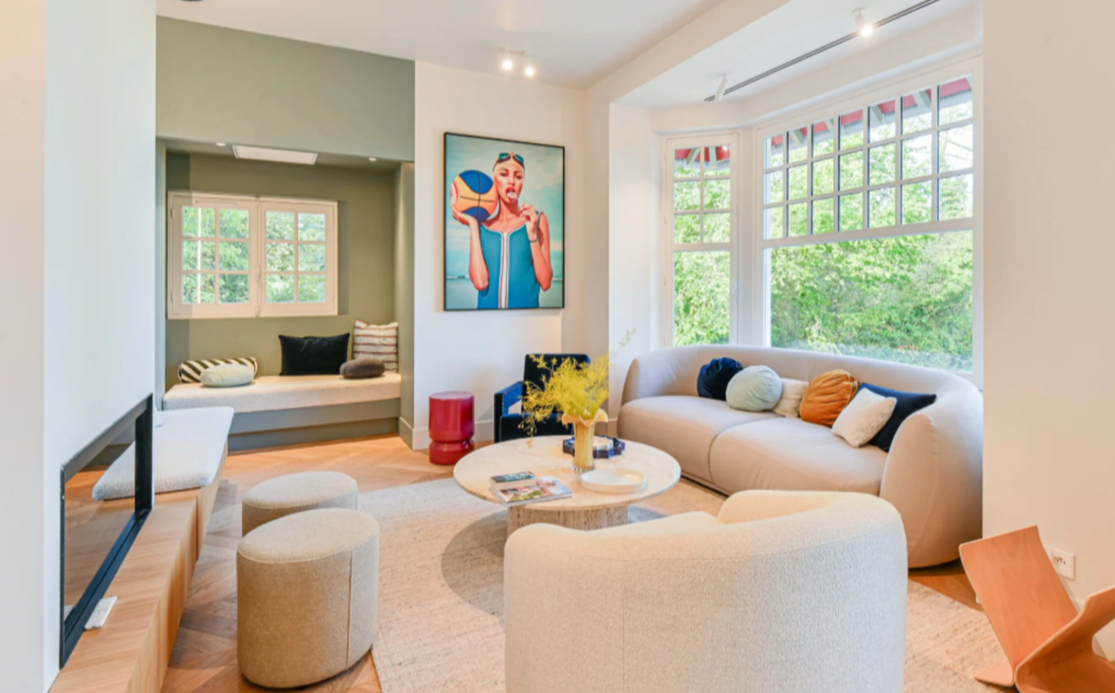 A modern living room features a curved sofa, round coffee table, and large windows, with a colorful artwork hanging on the wall.