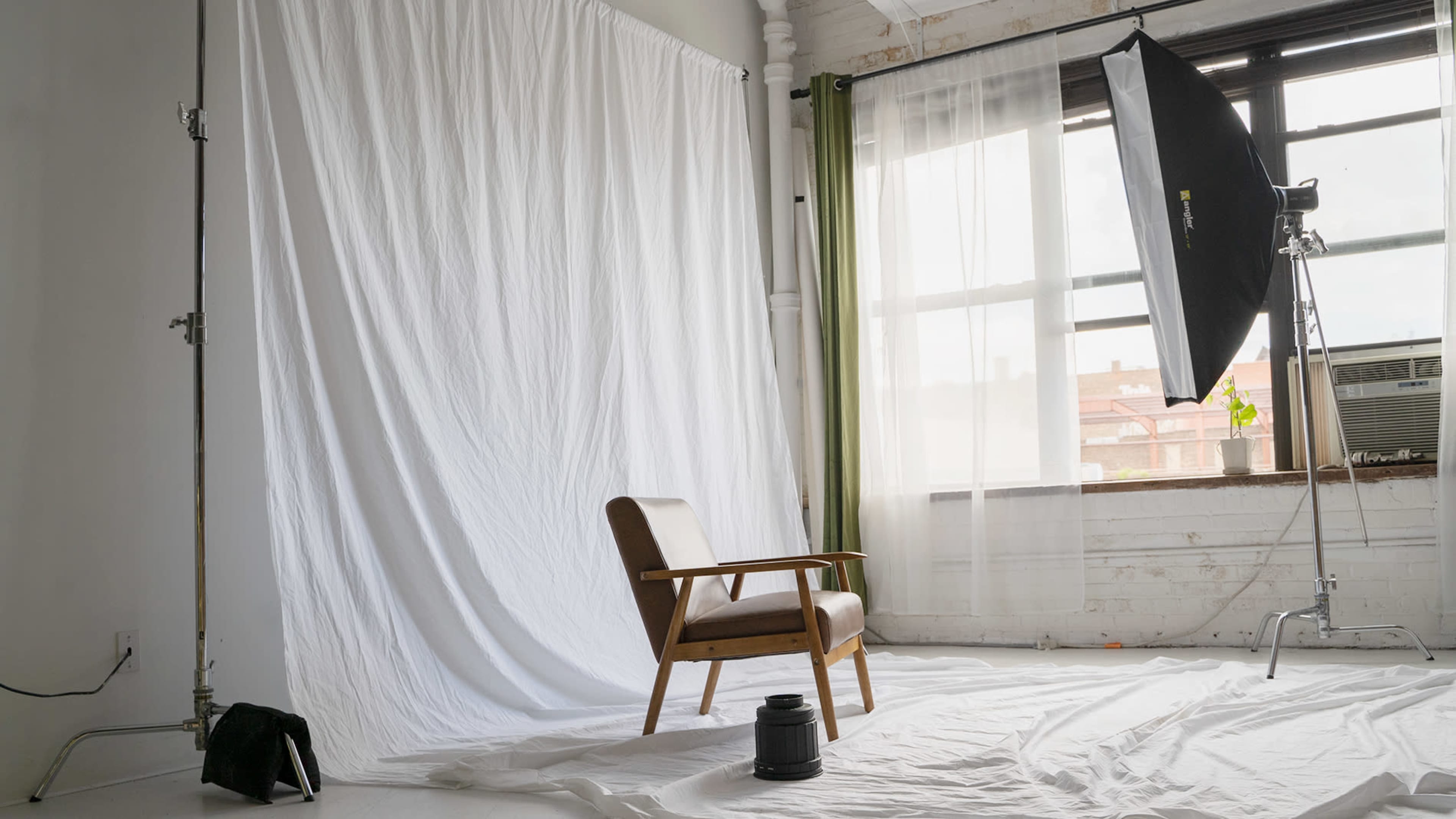 A wooden chair sits in front of a white backdrop in a well-lit photography studio with large windows.