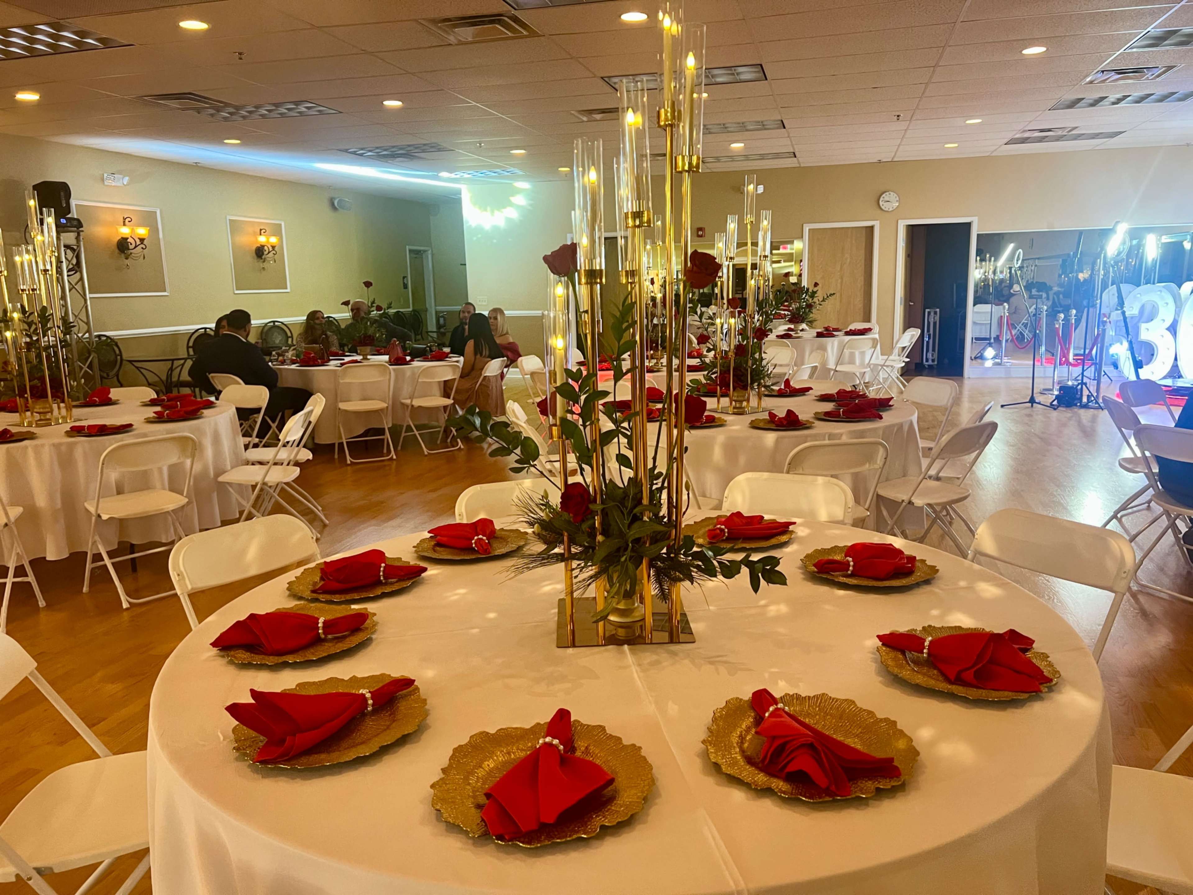 A banquet hall is set up with round tables adorned with white tablecloths, red napkins, and gold accents, while background lighting creates a festive atmosphere.
