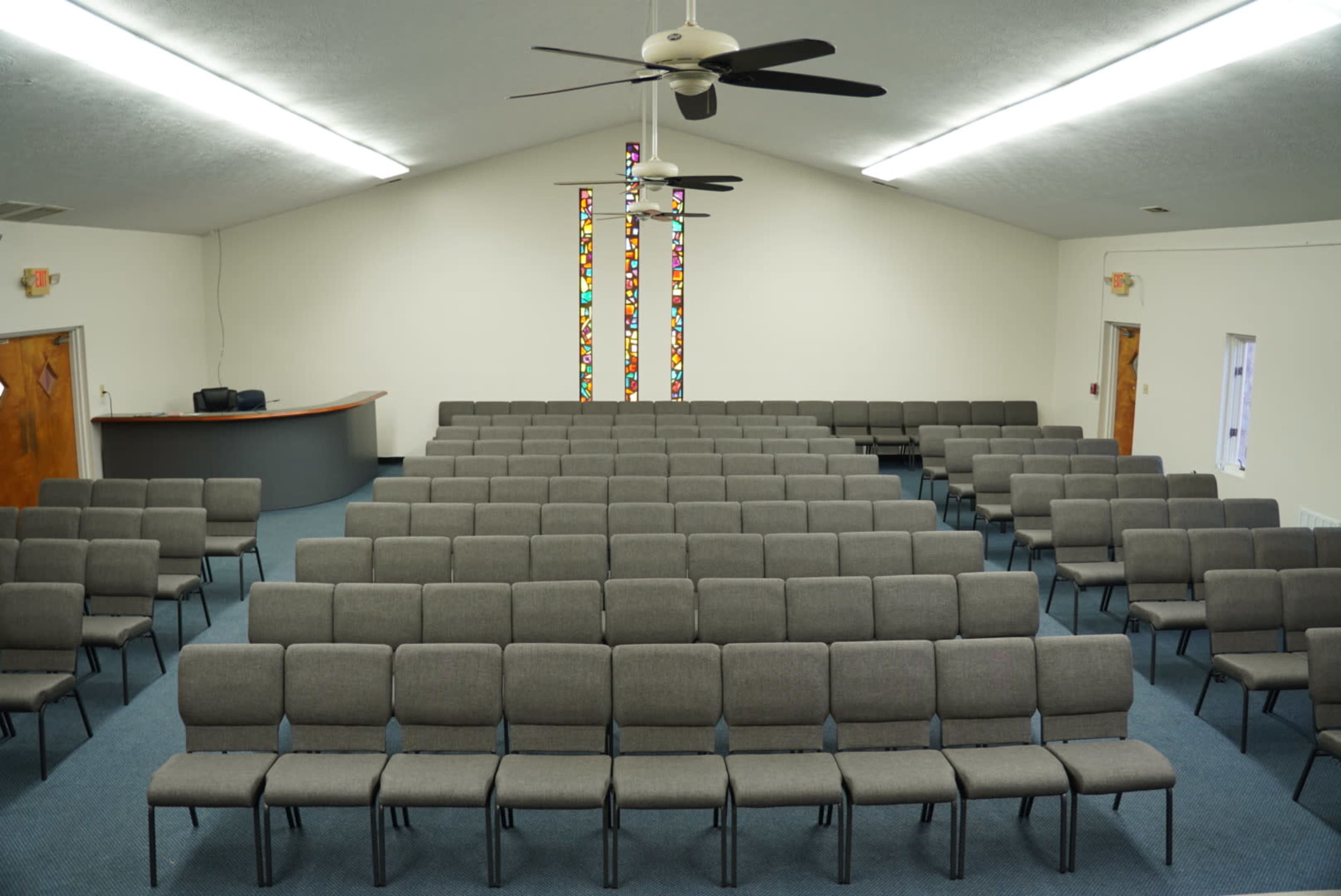 The image shows an empty auditorium with rows of gray chairs facing a stage area, adorned with stained glass windows.