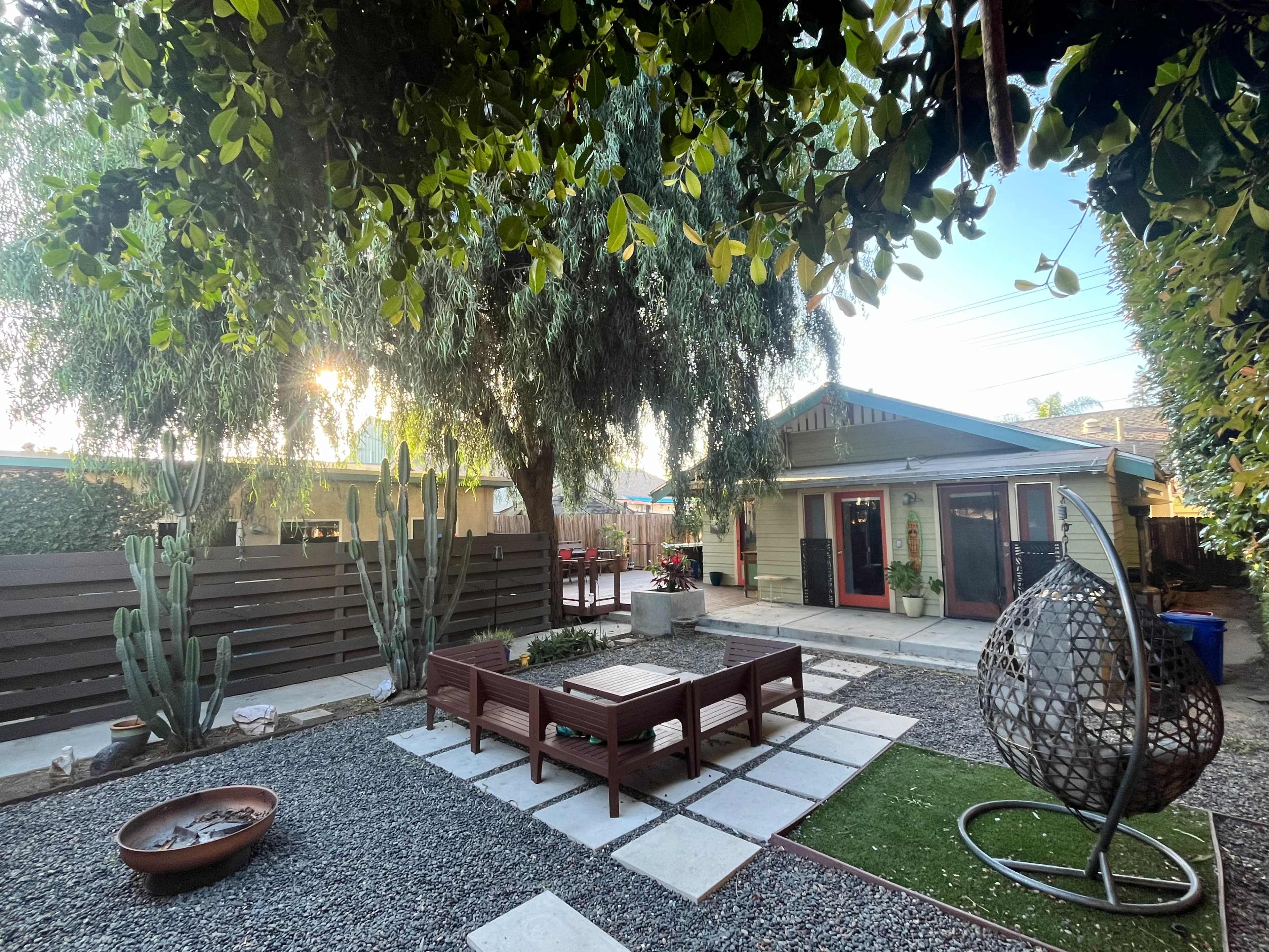 The image shows a landscaped backyard with a seating area, stone pathways, and a variety of plants, including cacti and trees, surrounding a small house.