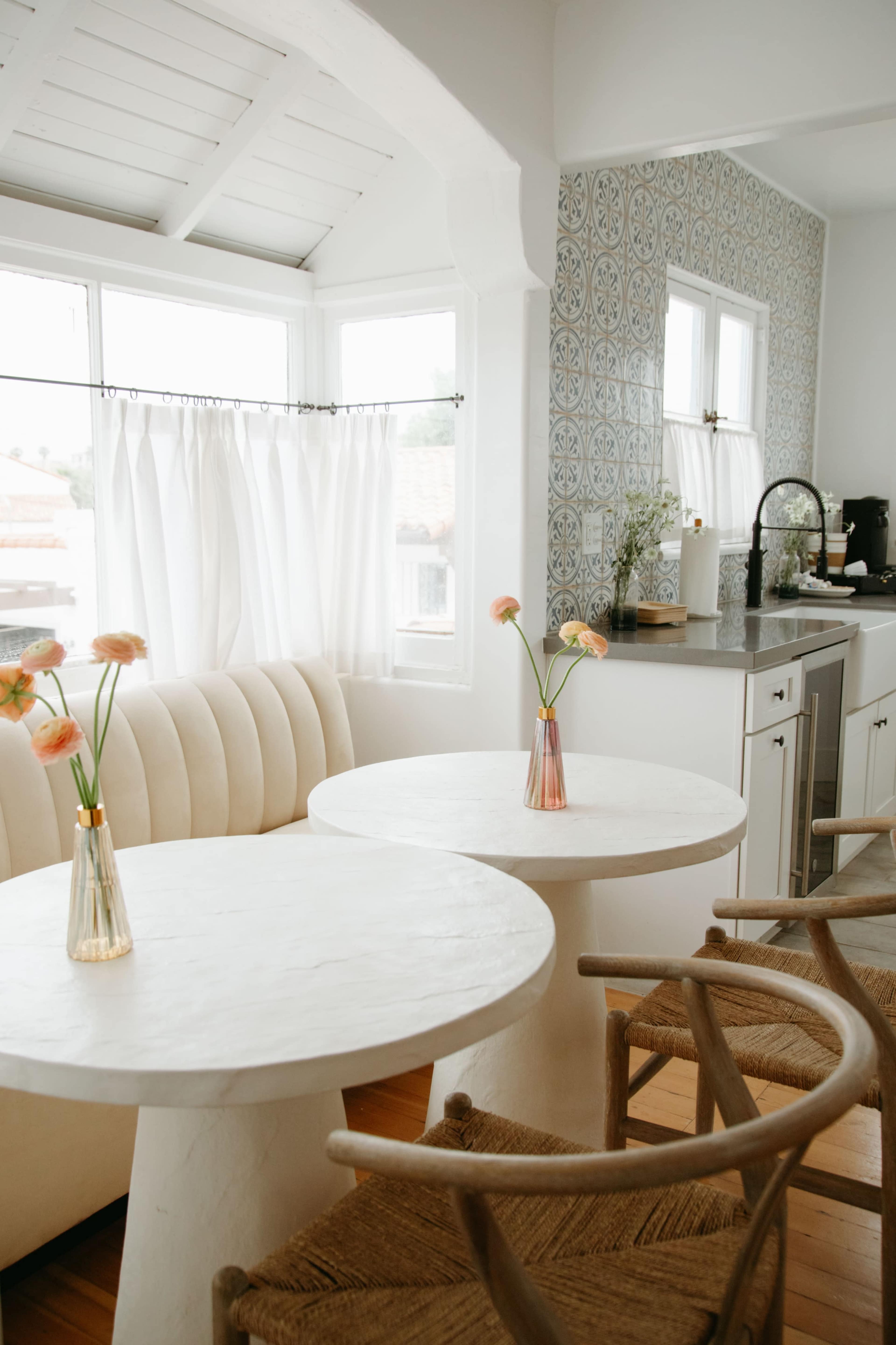 A bright kitchen with two white tables, each holding a vase of flowers, alongside a cozy seating area and patterned tile backsplash.
