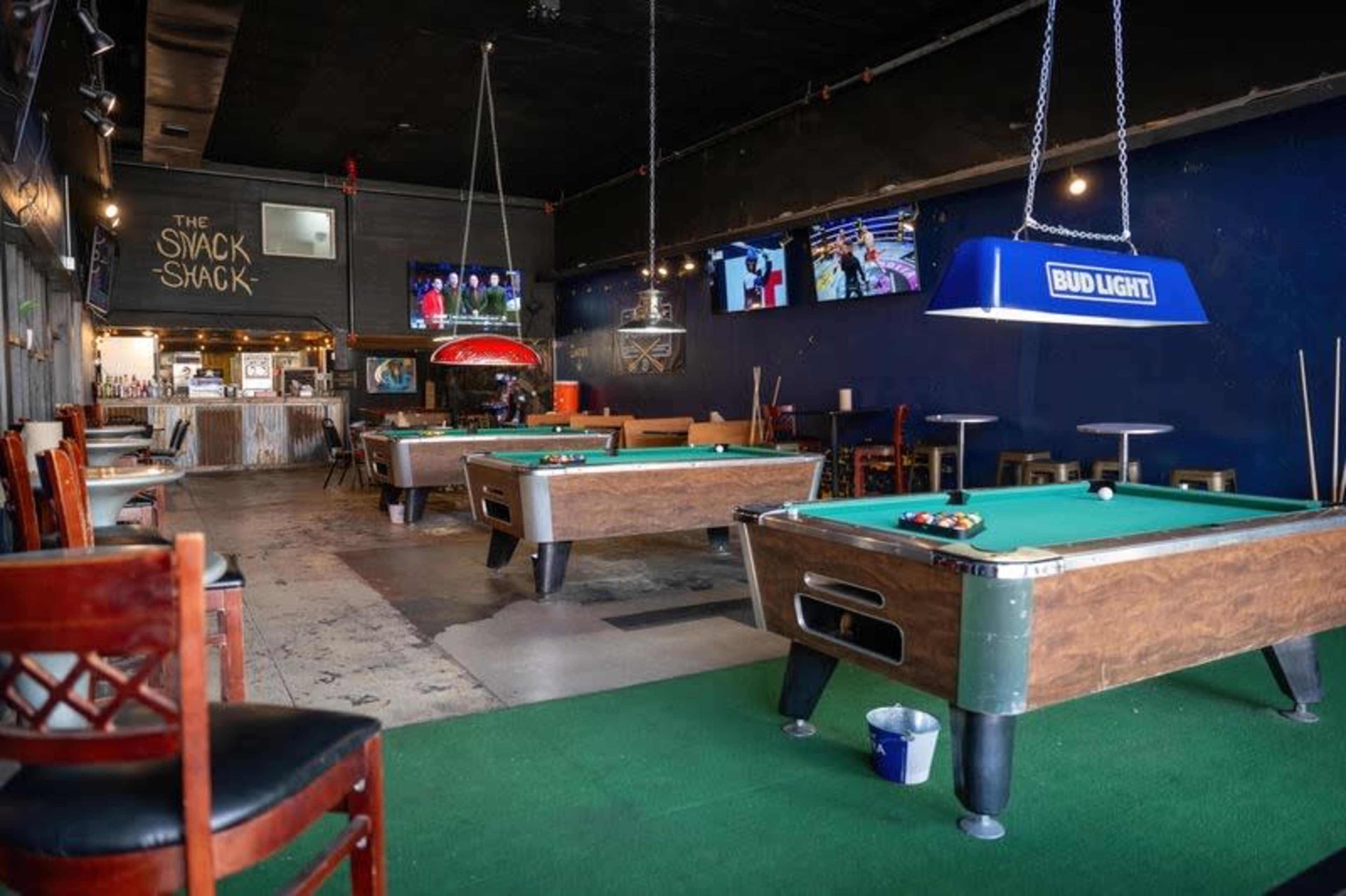 The image shows the interior of a bar with pool tables, a snack counter in the background, and several televisions on the walls.