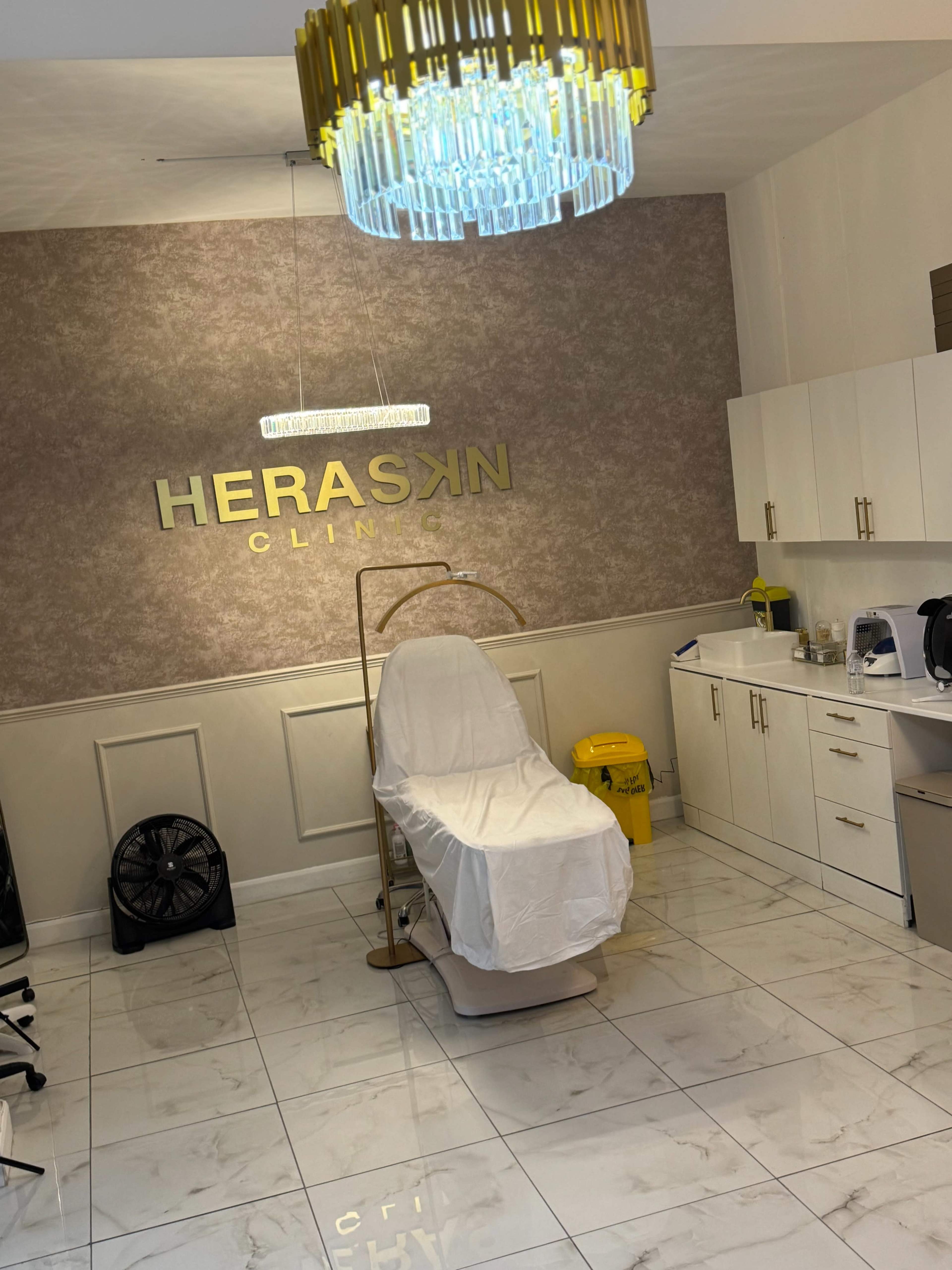 The image shows a clean clinic room with a treatment chair, cabinets, and a chandelier, featuring a sign that reads "HERASKIN CLINIC."