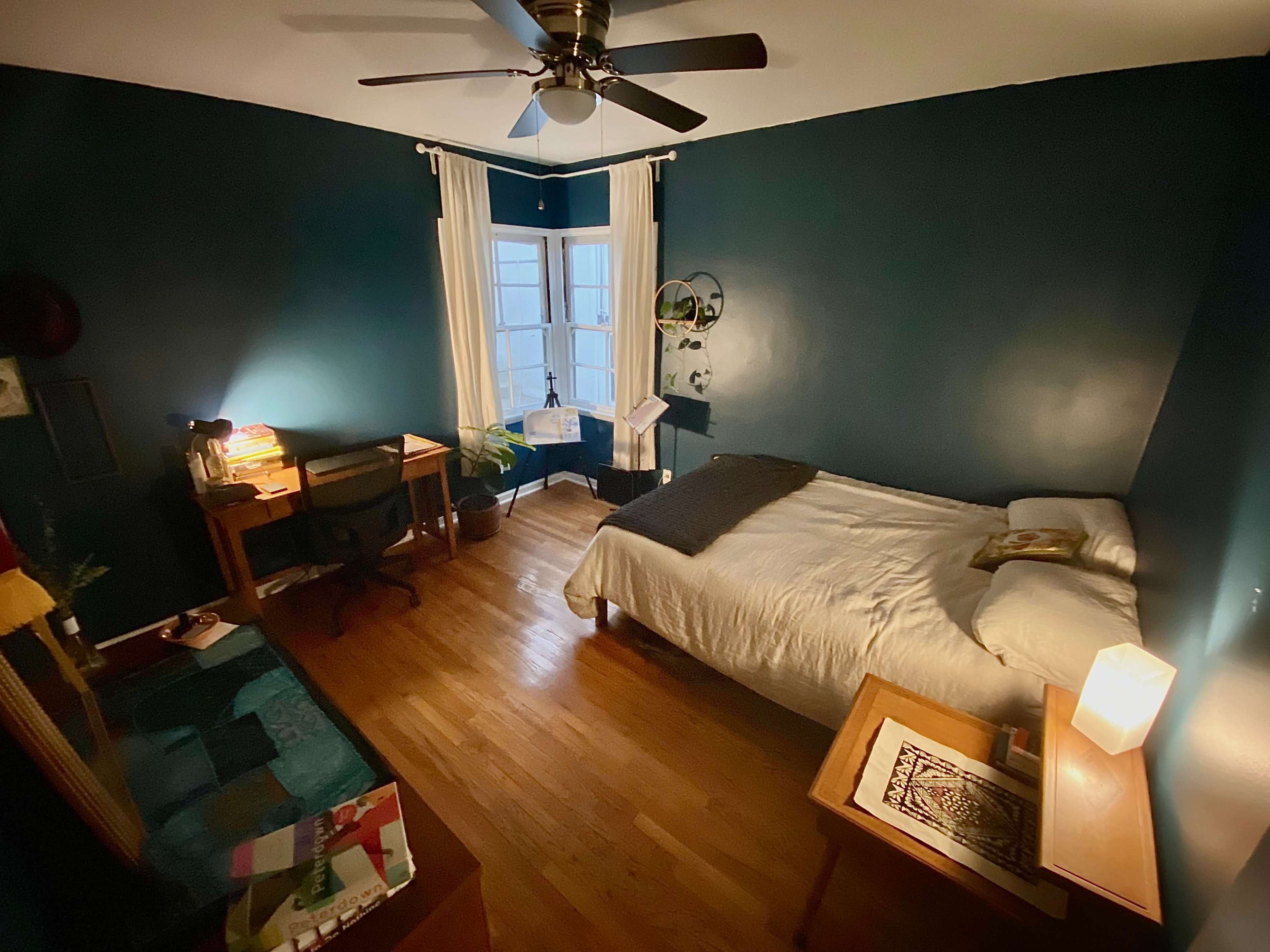 The image shows a cozy bedroom featuring a bed, a desk with a chair, and a window, all set against dark blue walls and wooden flooring.