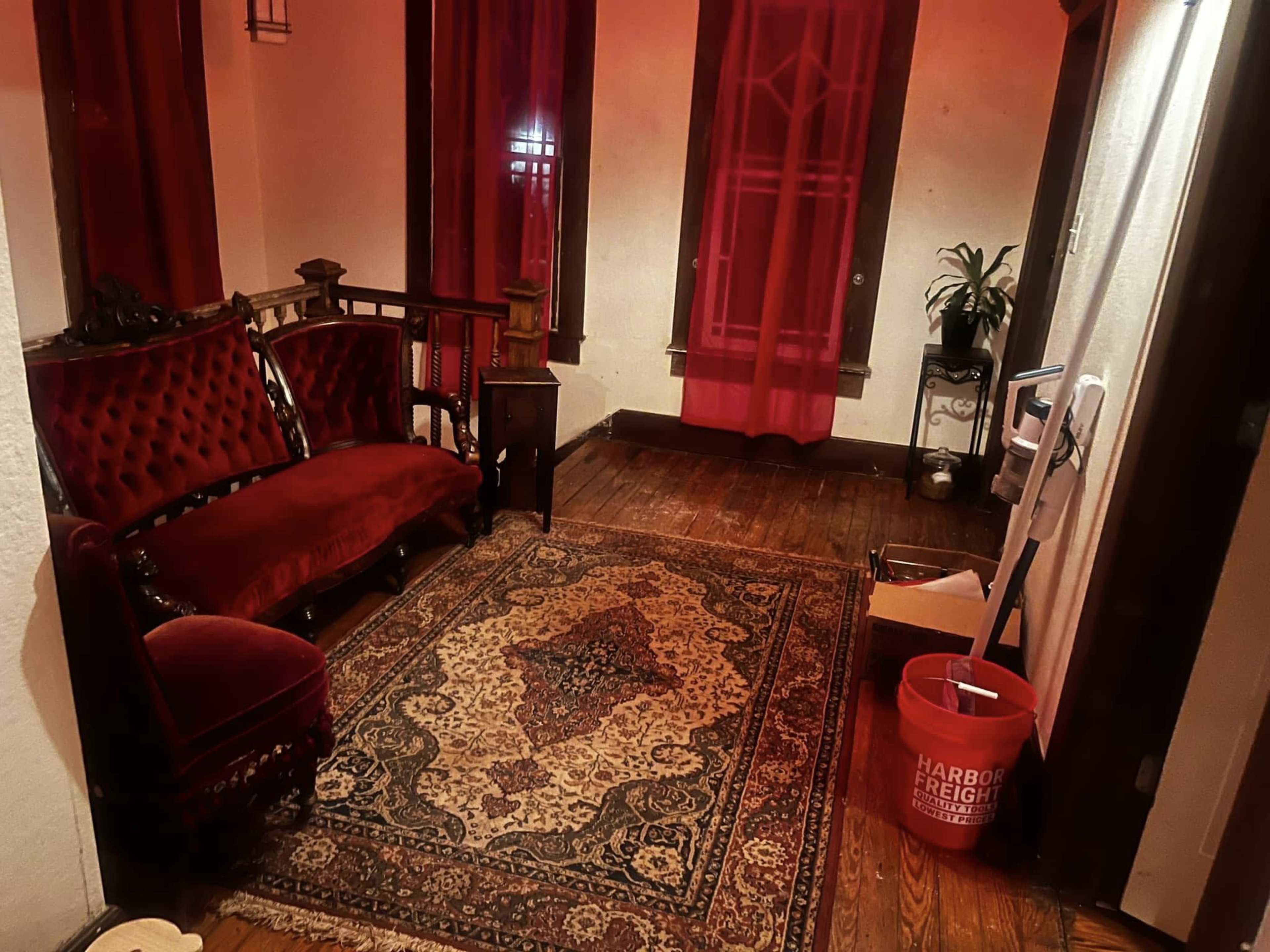 The image shows a dimly lit room with red curtains, a vintage red velvet sofa, a rug, and a small table beside the sofa.