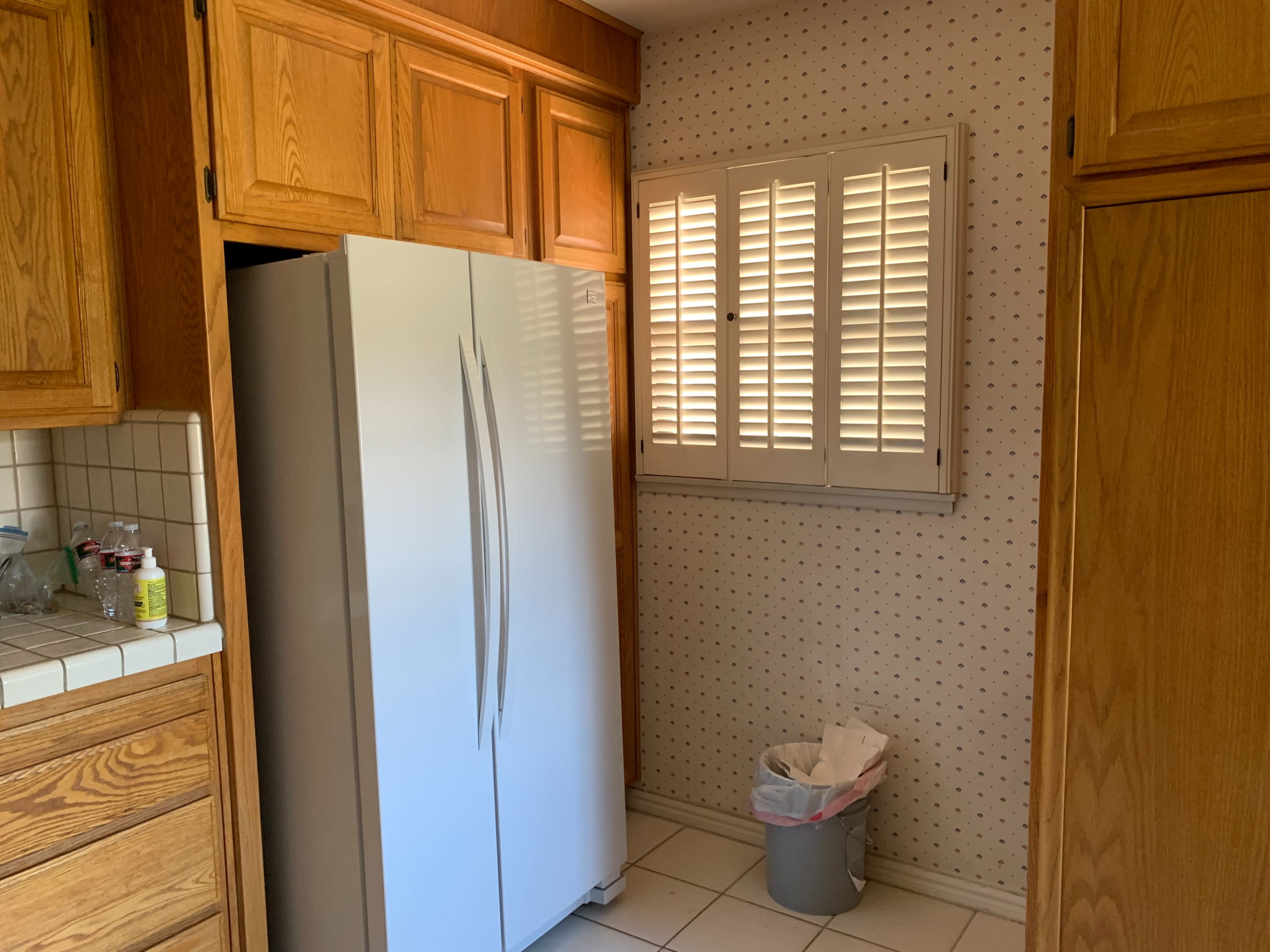 A white refrigerator stands next to wooden cabinets in a kitchen with a window covered by slatted shutters and a trash can on the floor.