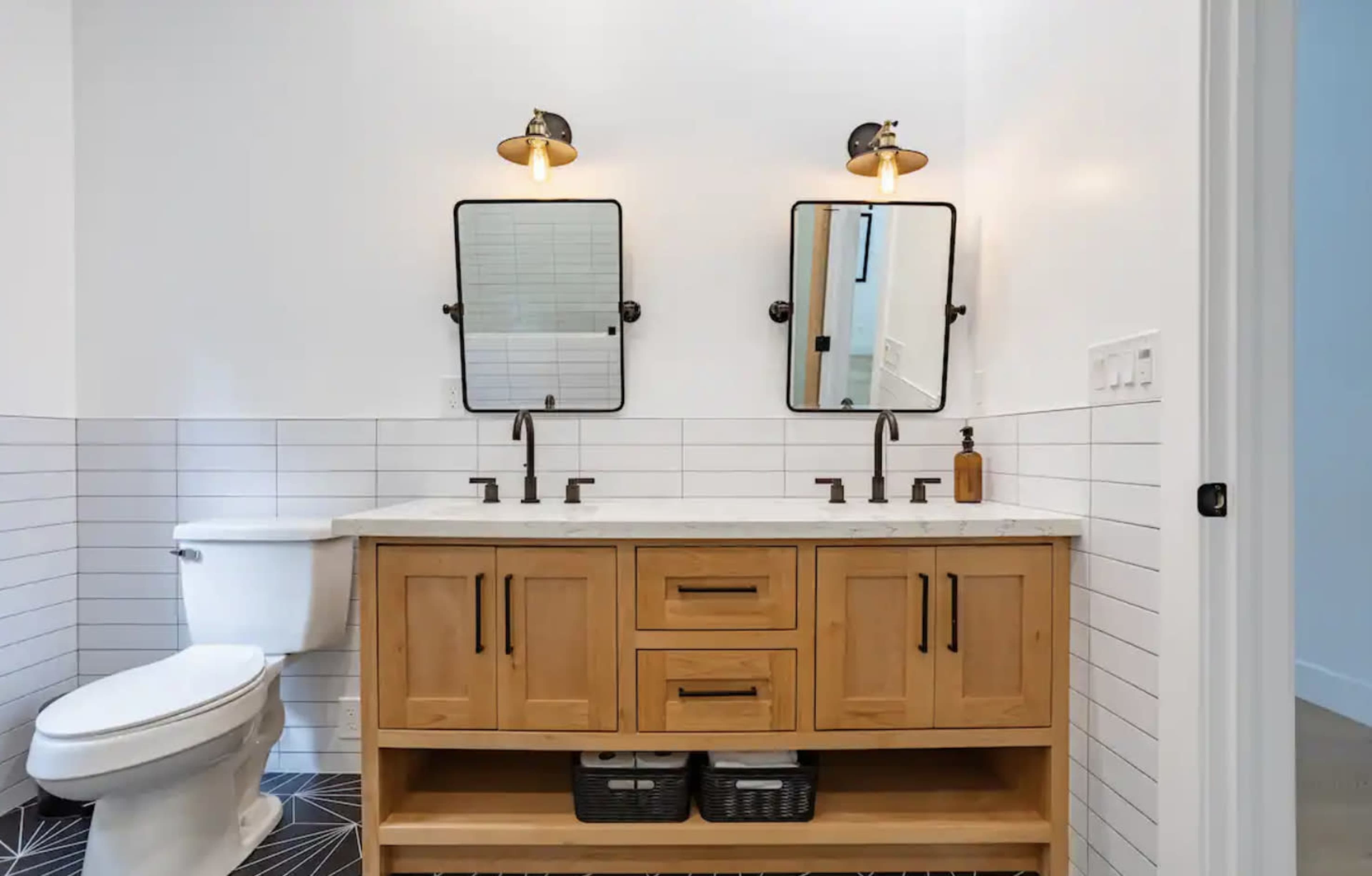 A modern bathroom featuring a wooden vanity with two sinks, two mirrors, and a toilet, all complemented by black fixtures and a tiled floor.