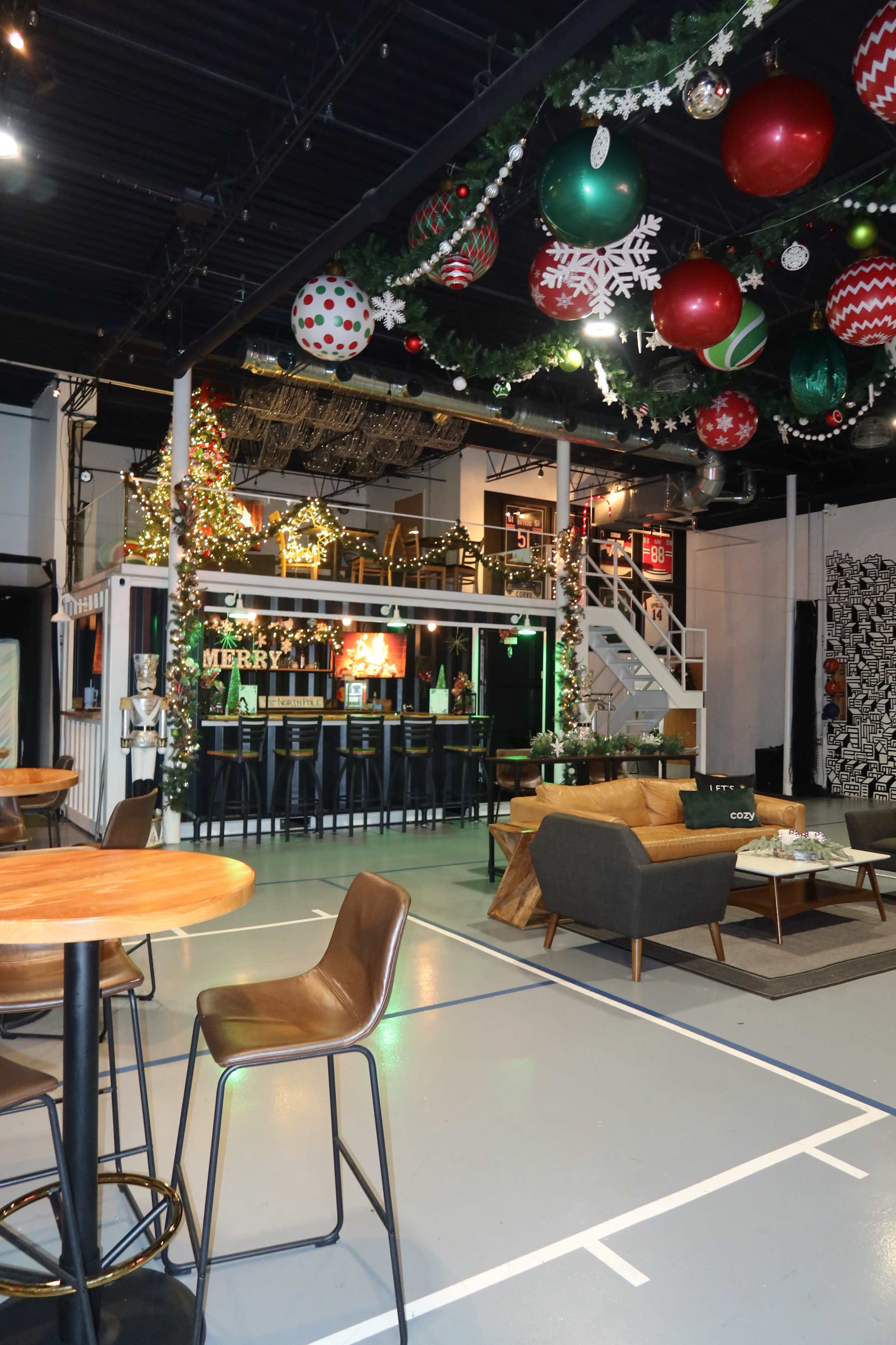 The image shows a decorated indoor space with festive holiday ornaments hanging from the ceiling, including large red and green balls, and a bar area in the background with Christmas decor.