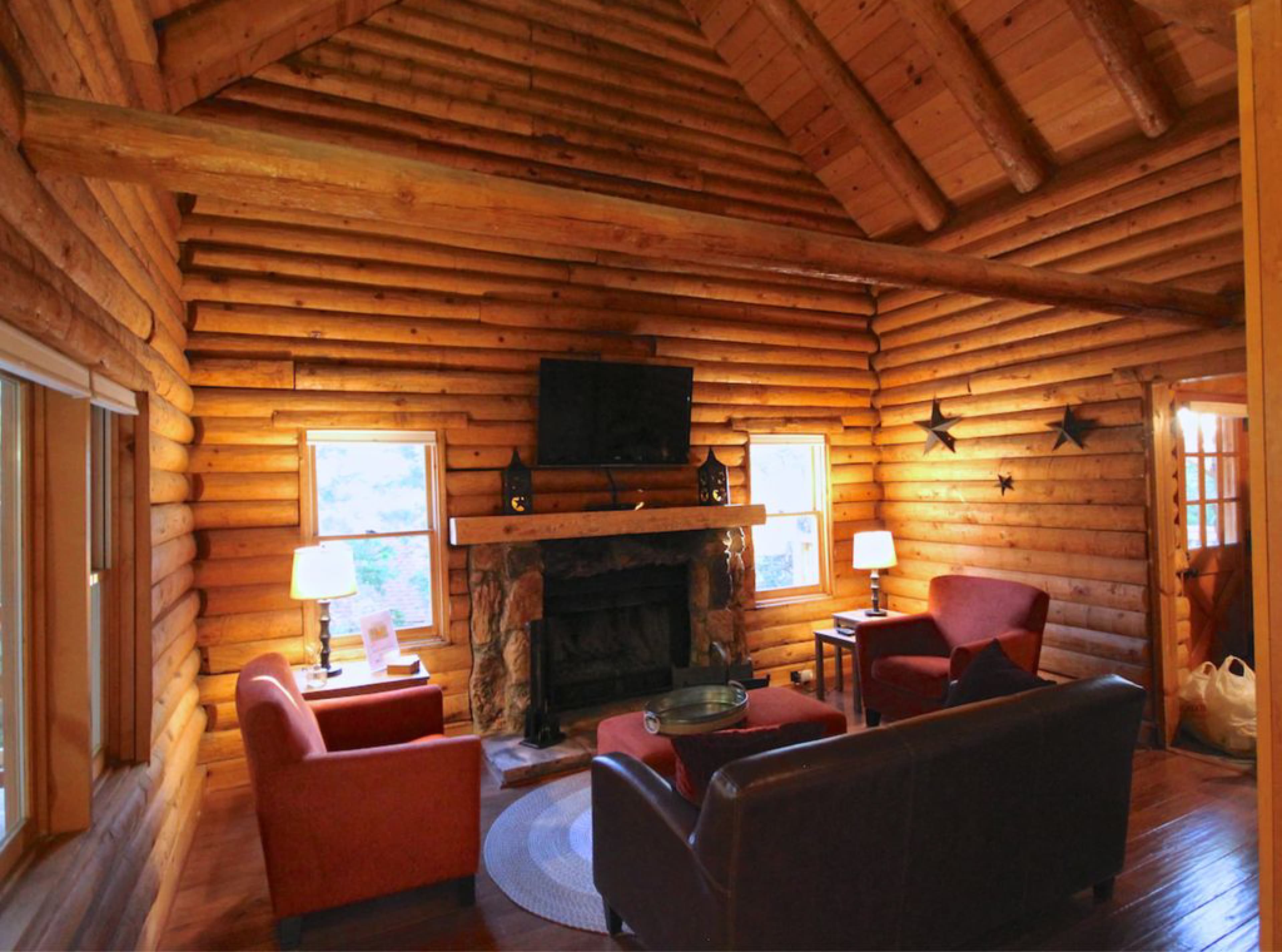 The interior of a log cabin features a cozy seating arrangement with two red armchairs and a dark sofa, a stone fireplace, and large windows allowing natural light.