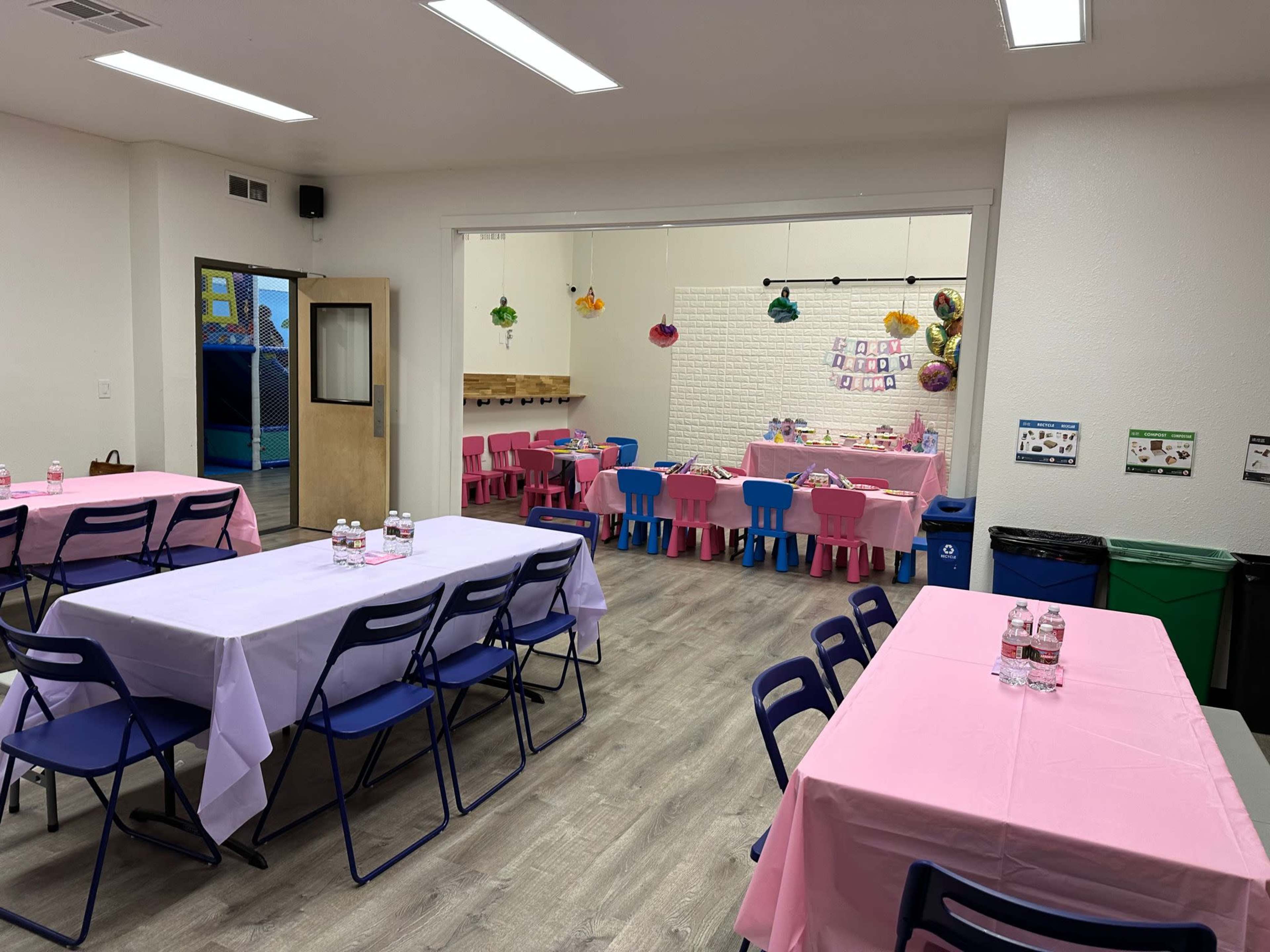 A room with several tables covered in pink and white tablecloths, separated by blue chairs, with a play area visible in the background.