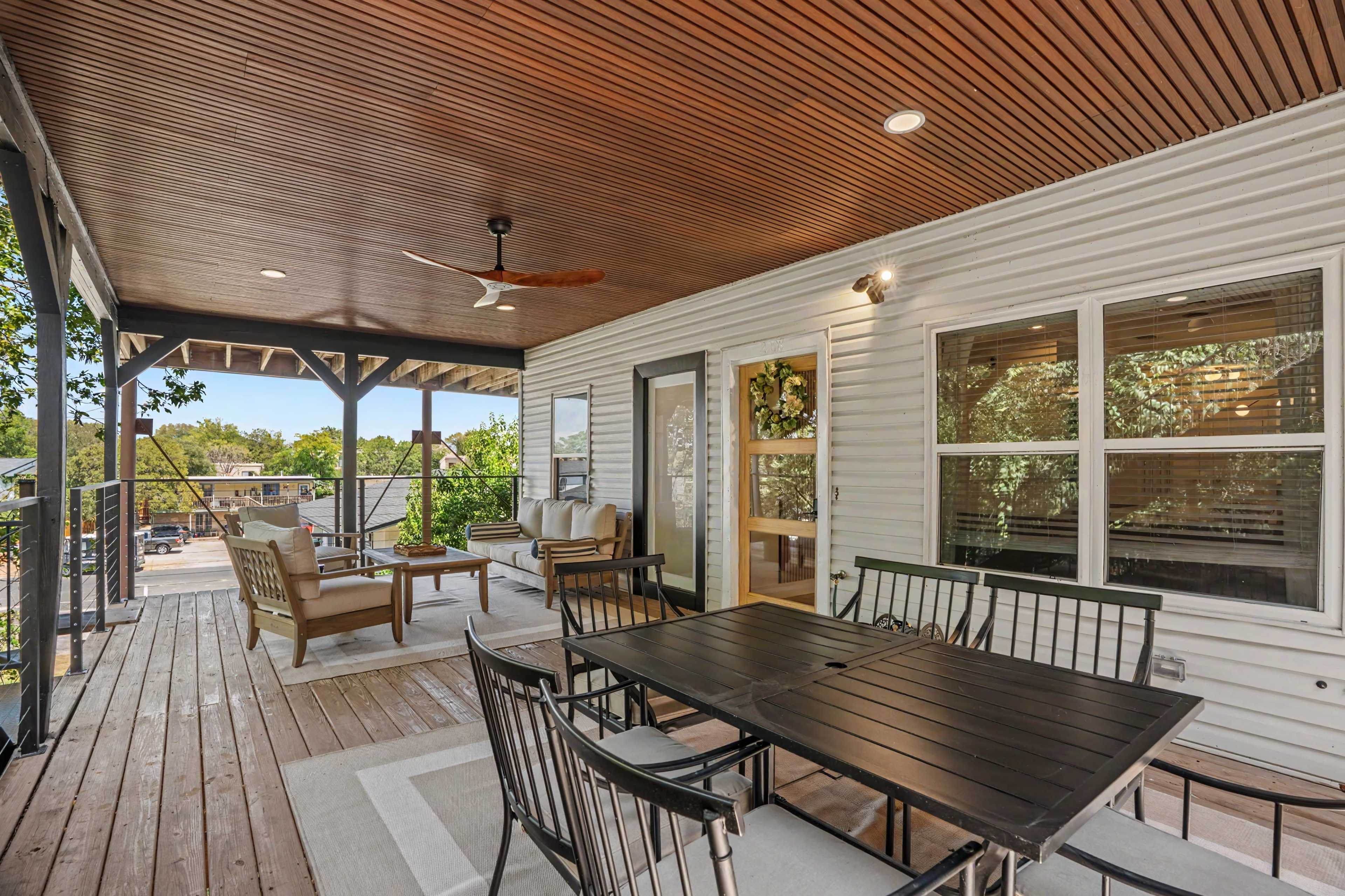 The image shows a spacious outdoor patio area featuring a dining table with chairs, a seating arrangement, a ceiling fan, and wooden decking.