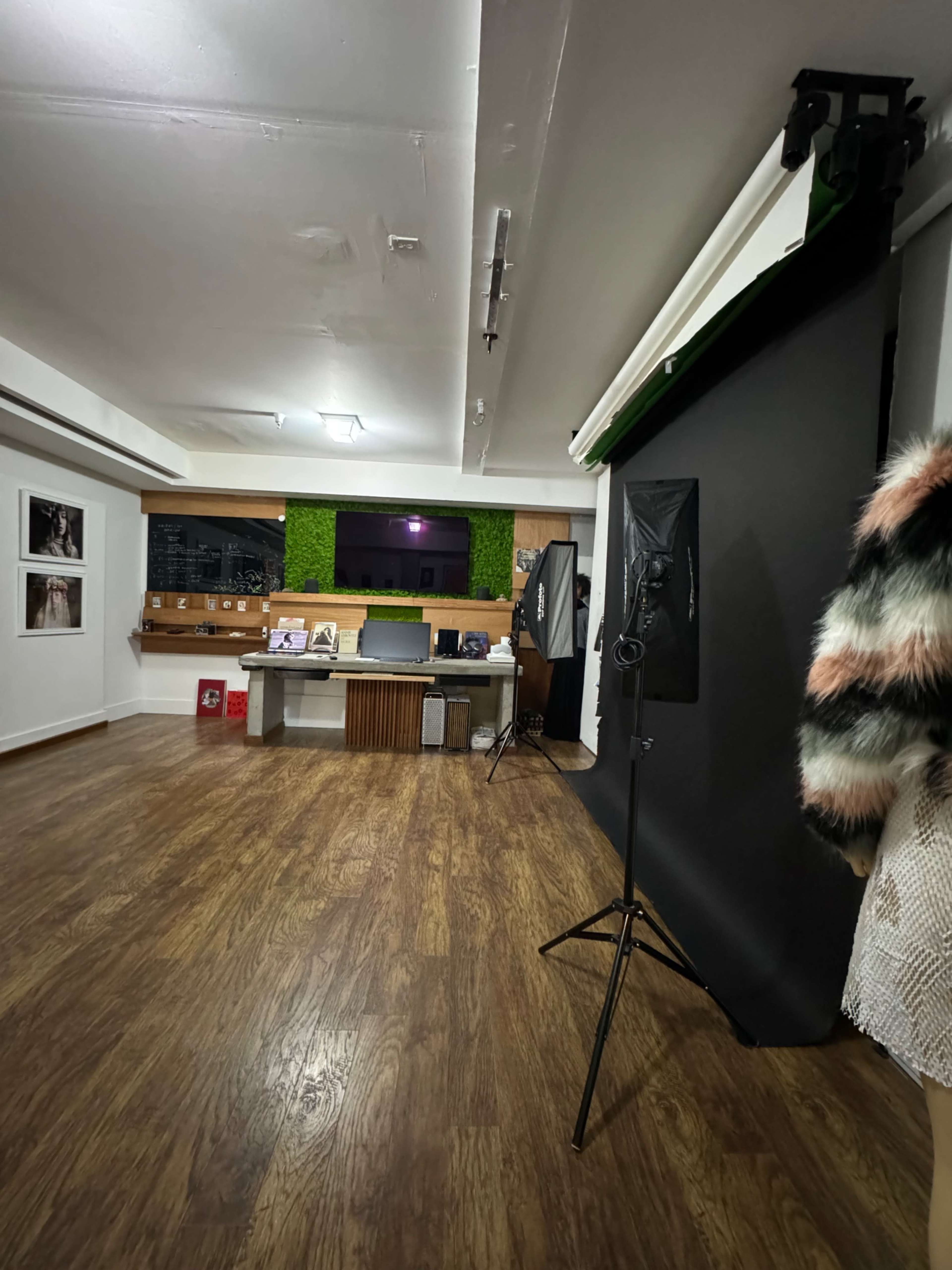 A spacious interior with wooden flooring, a dark backdrop, softbox lighting equipment, and a small reception area featuring a green wall and a desk.