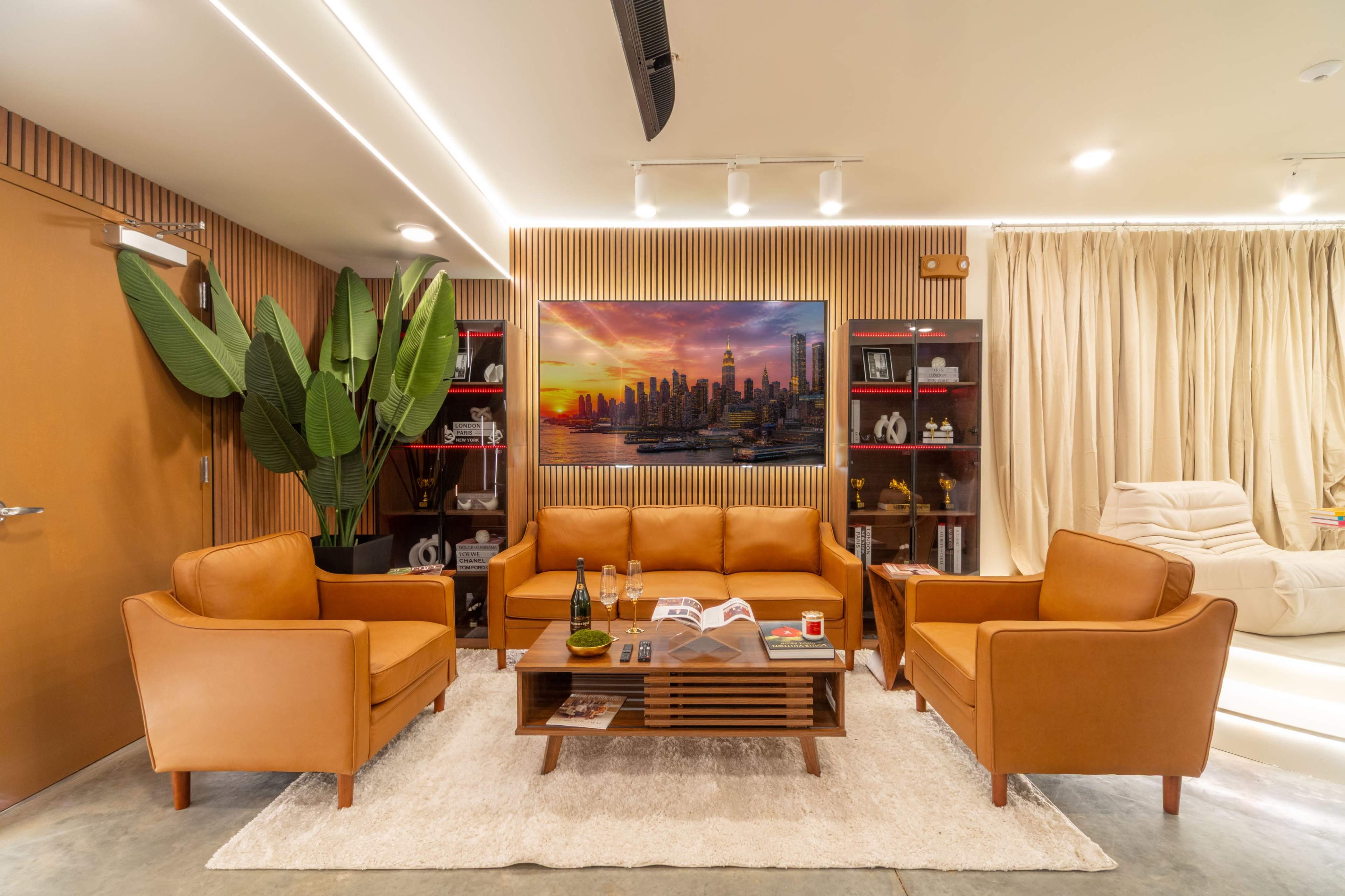 The image shows a modern living room featuring a brown leather sofa and armchairs, a wooden coffee table, and a large painting of a city skyline on the wall.