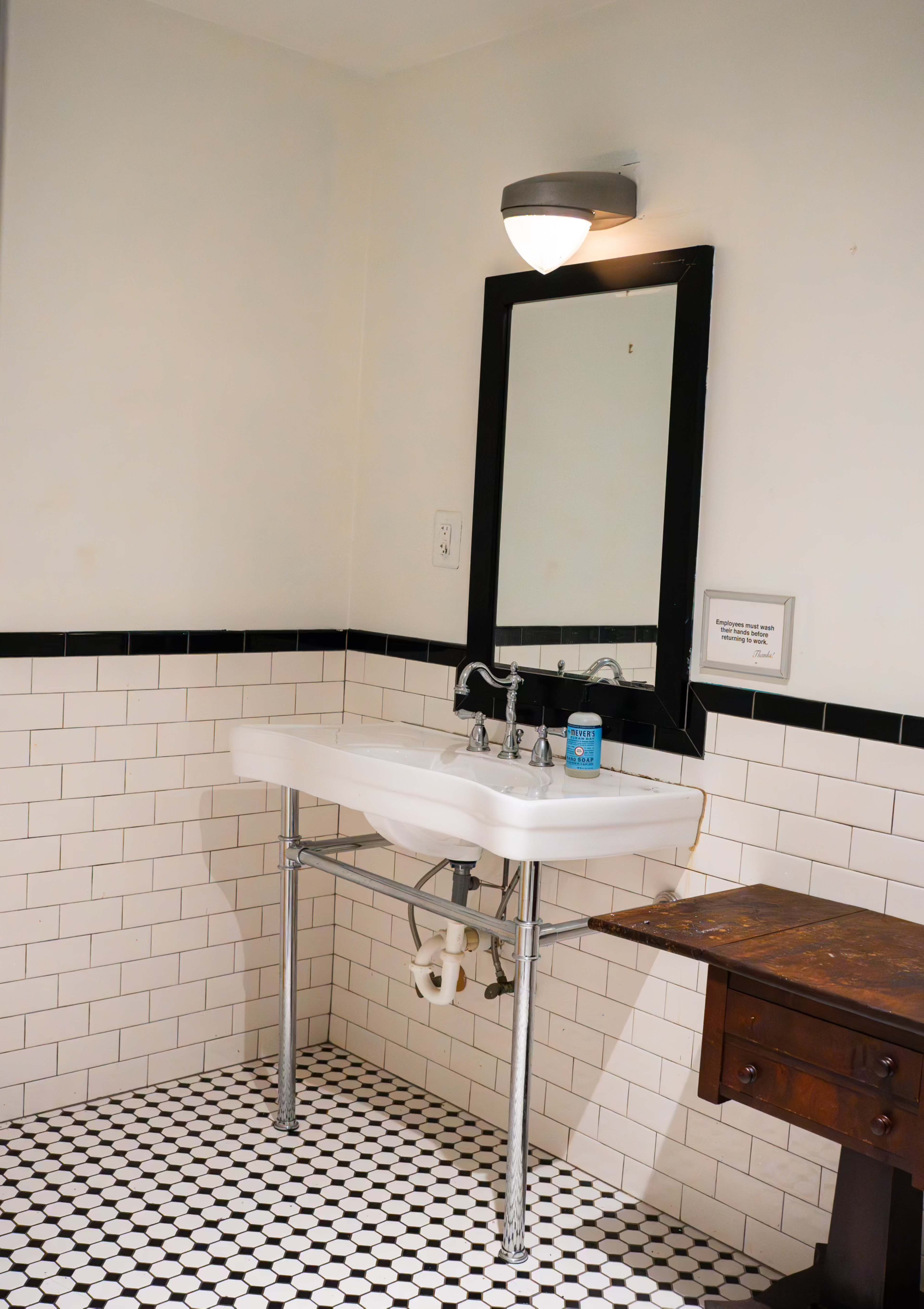 A bathroom with a white pedestal sink, a mirror above it, and a wooden side table on the right, all set against a tiled floor with black and white patterns.