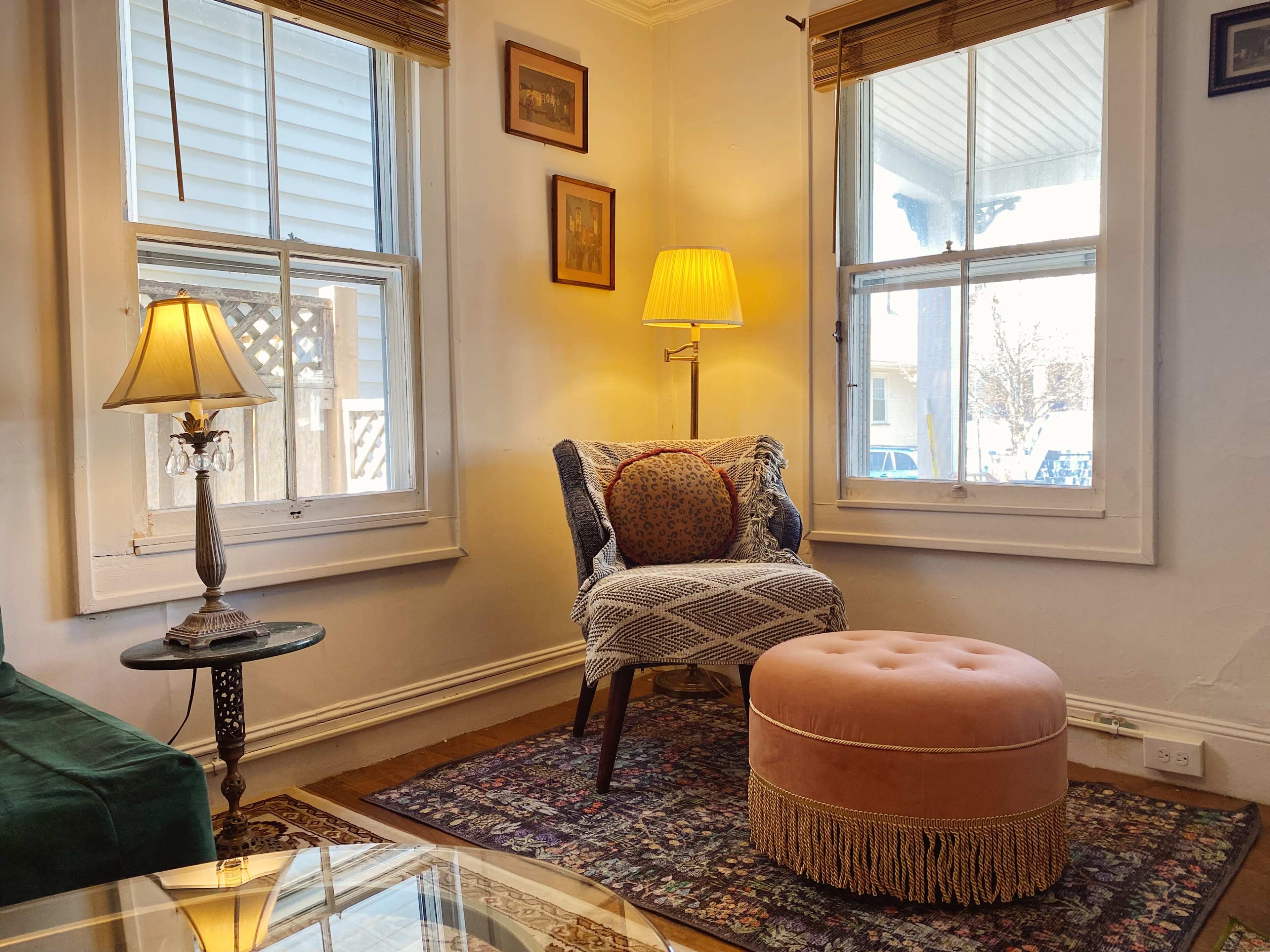 A cozy reading nook features an armchair with a decorative pillow, a round ottoman, and a nearby table lamp, all positioned by large windows.