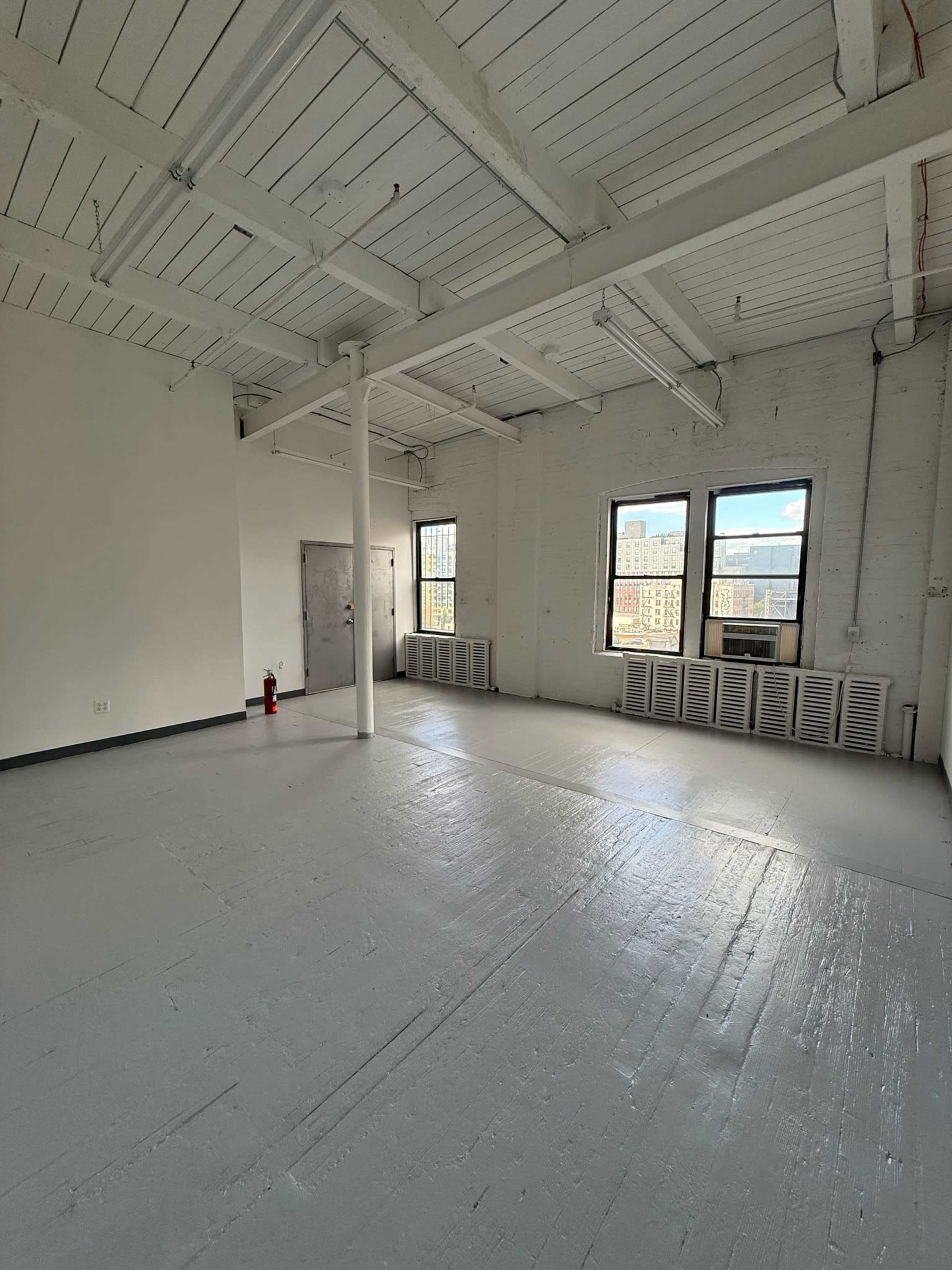 The image depicts a spacious, brightly lit room with white walls, a polished floor, and large windows.