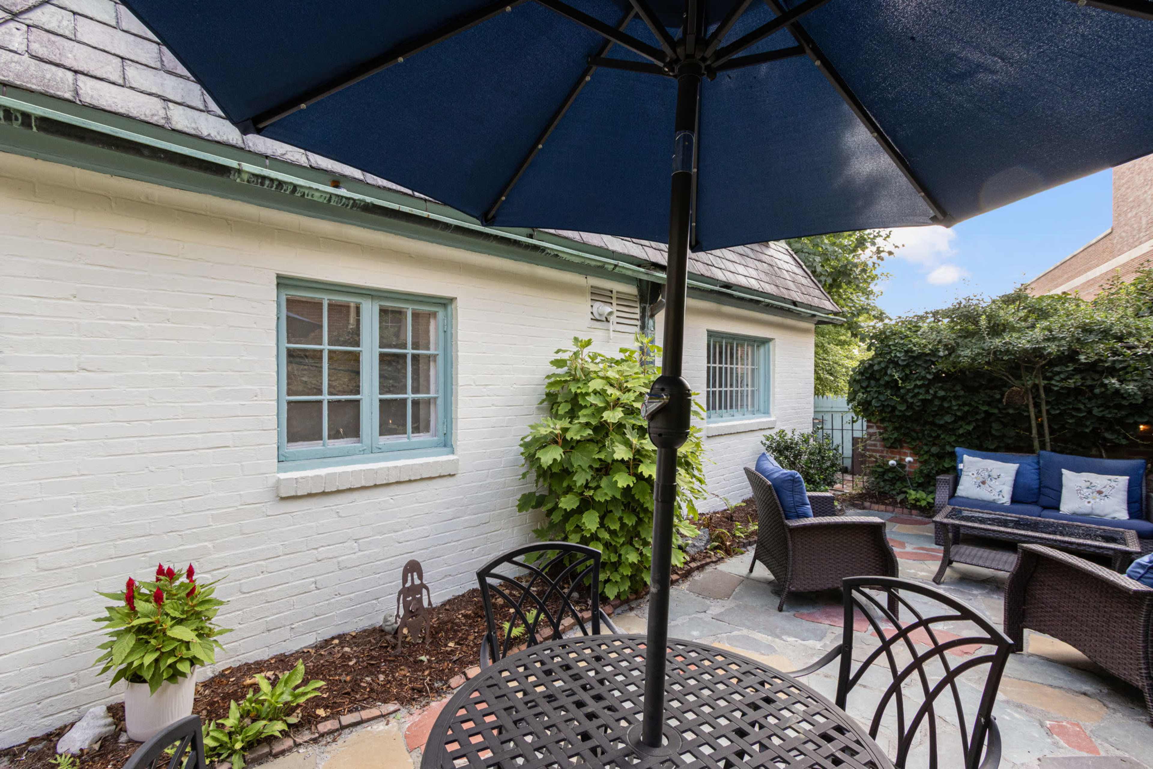 A small outdoor patio features a round table with black chairs, an umbrella, and a seating area surrounded by greenery and a brick wall.