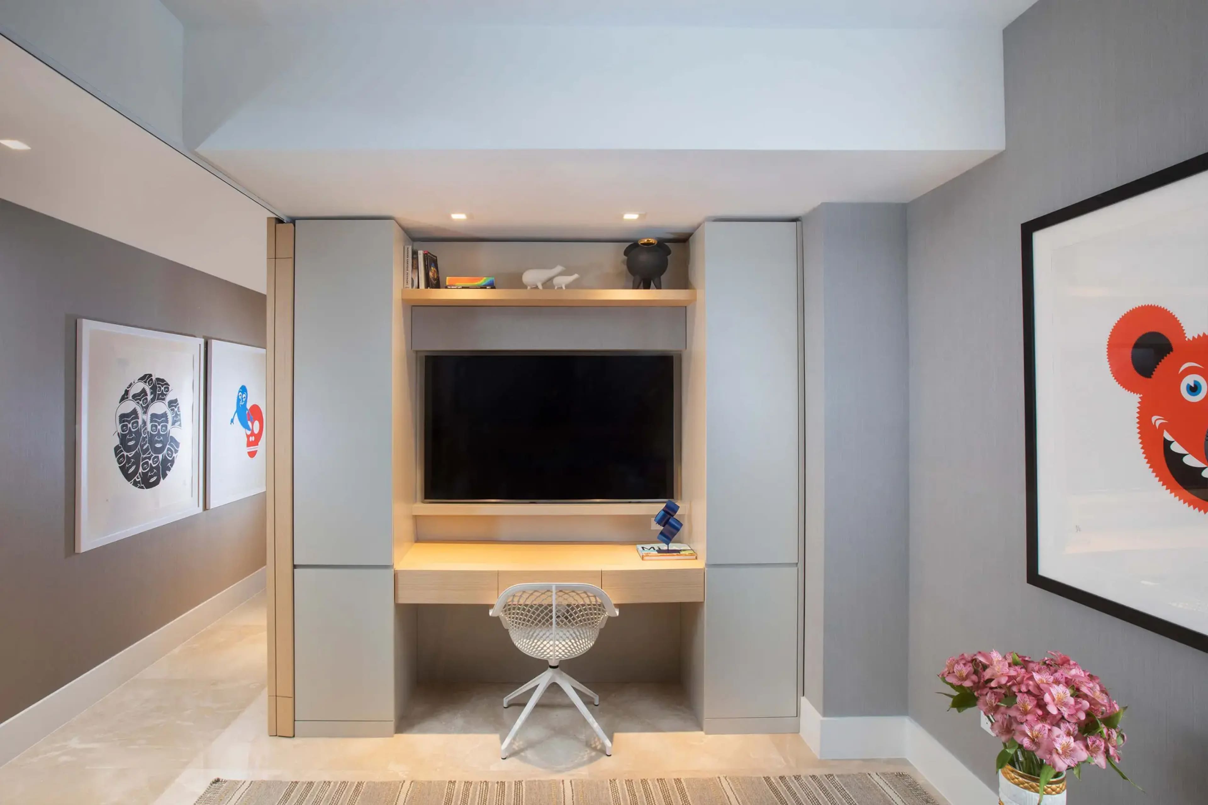 A modern workspace featuring a wall-mounted television, a light wooden desk with a white chair, and artwork displayed on the adjacent walls.