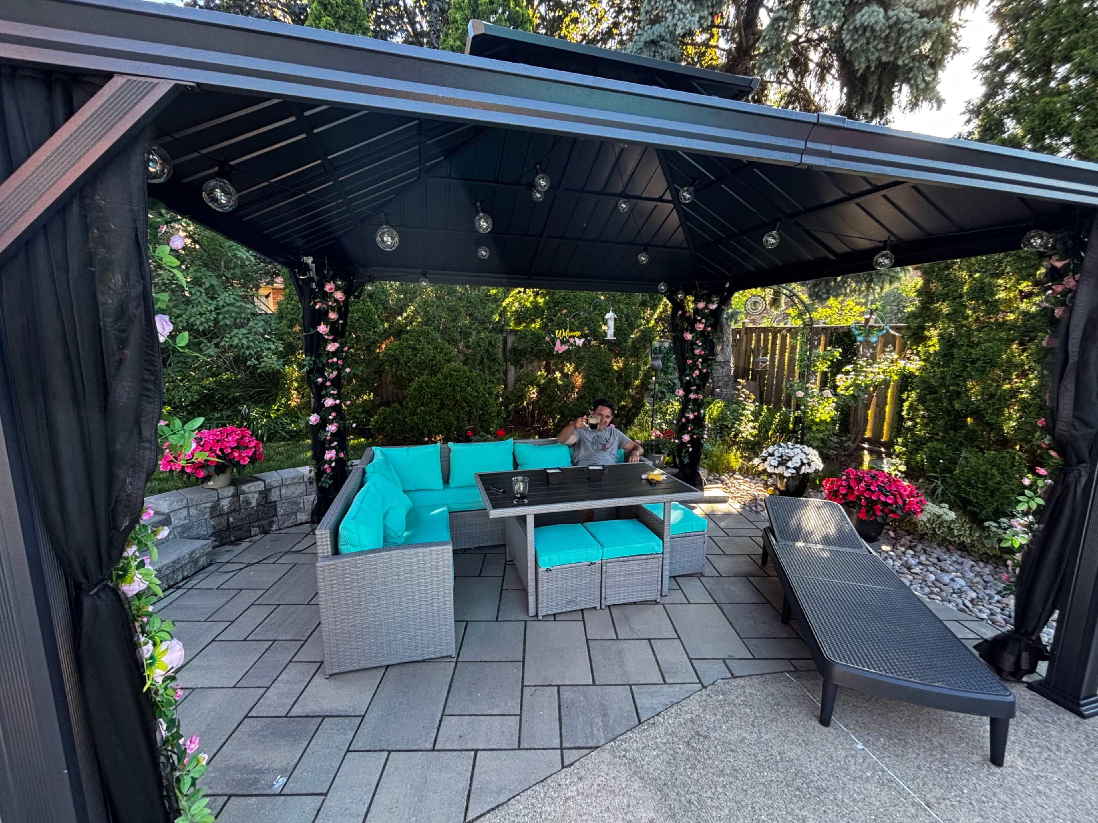 A person sits on a turquoise outdoor sofa under a gazebo surrounded by lush greenery and colorful flowers.