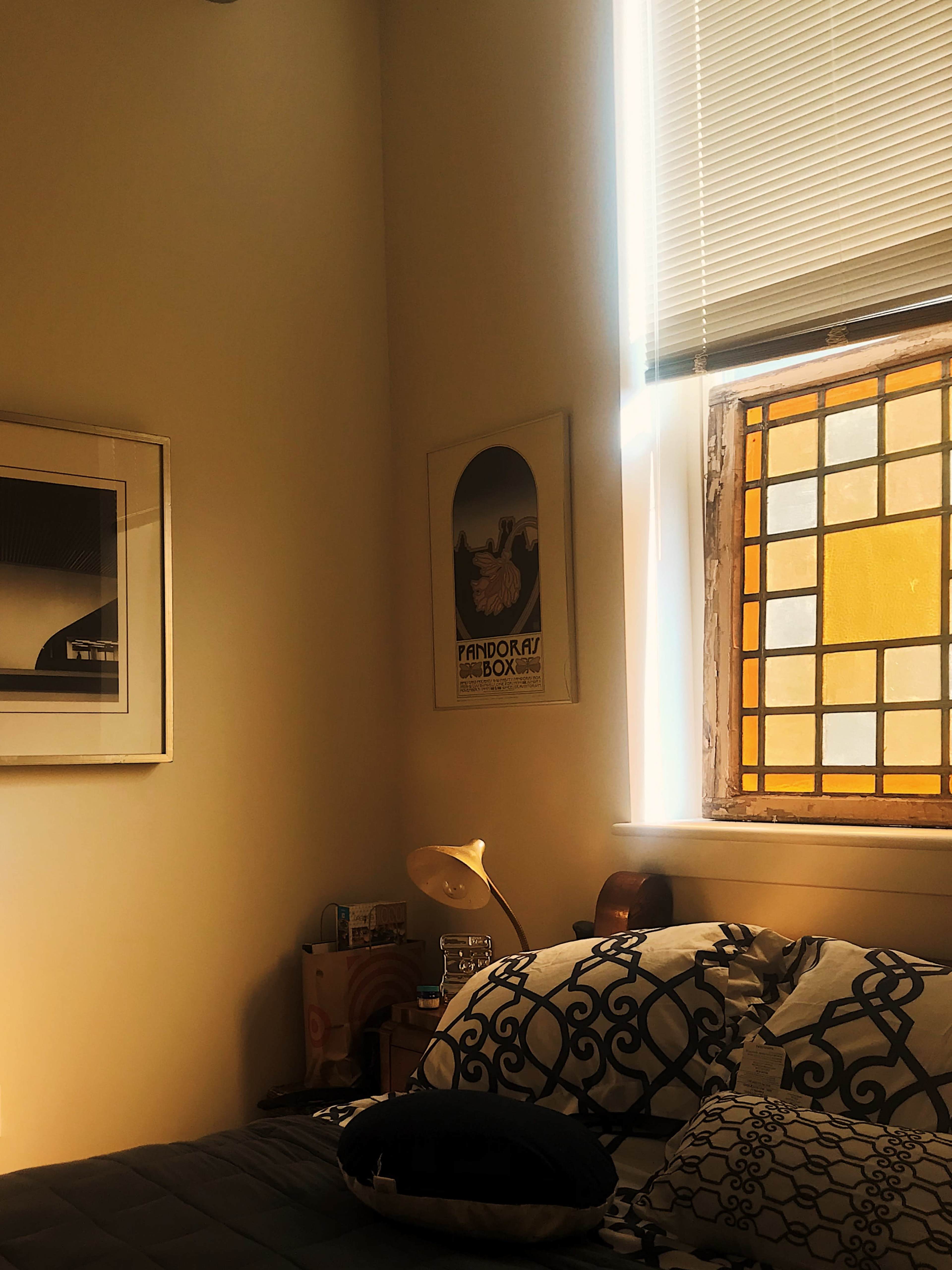 A softly lit bedroom featuring a bed with patterned pillows, a framed artwork on the wall, and a window with stained glass.