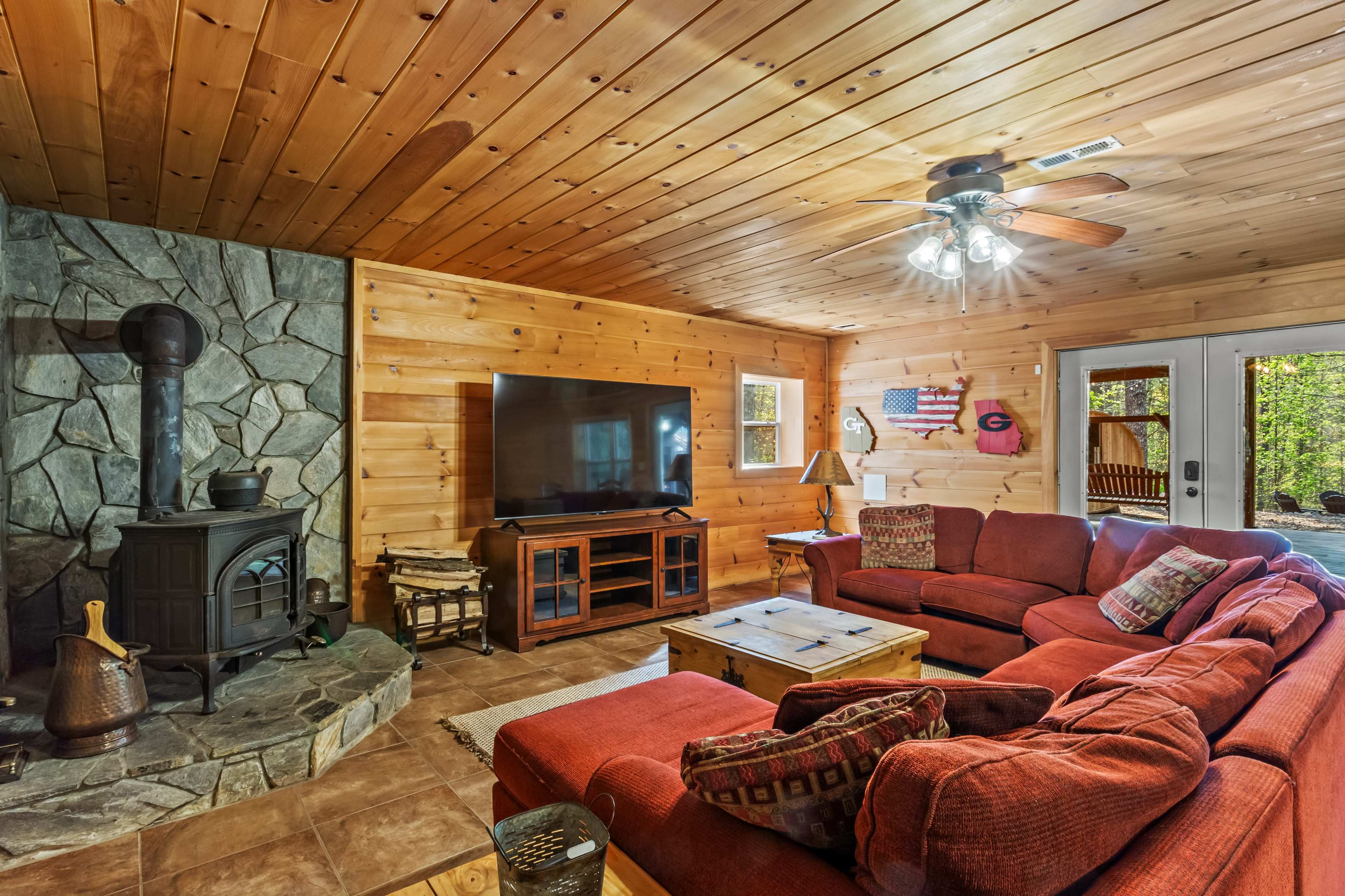 A cozy wooden cabin living room features a stone fireplace, a large television, and a sectional sofa set around a coffee table.