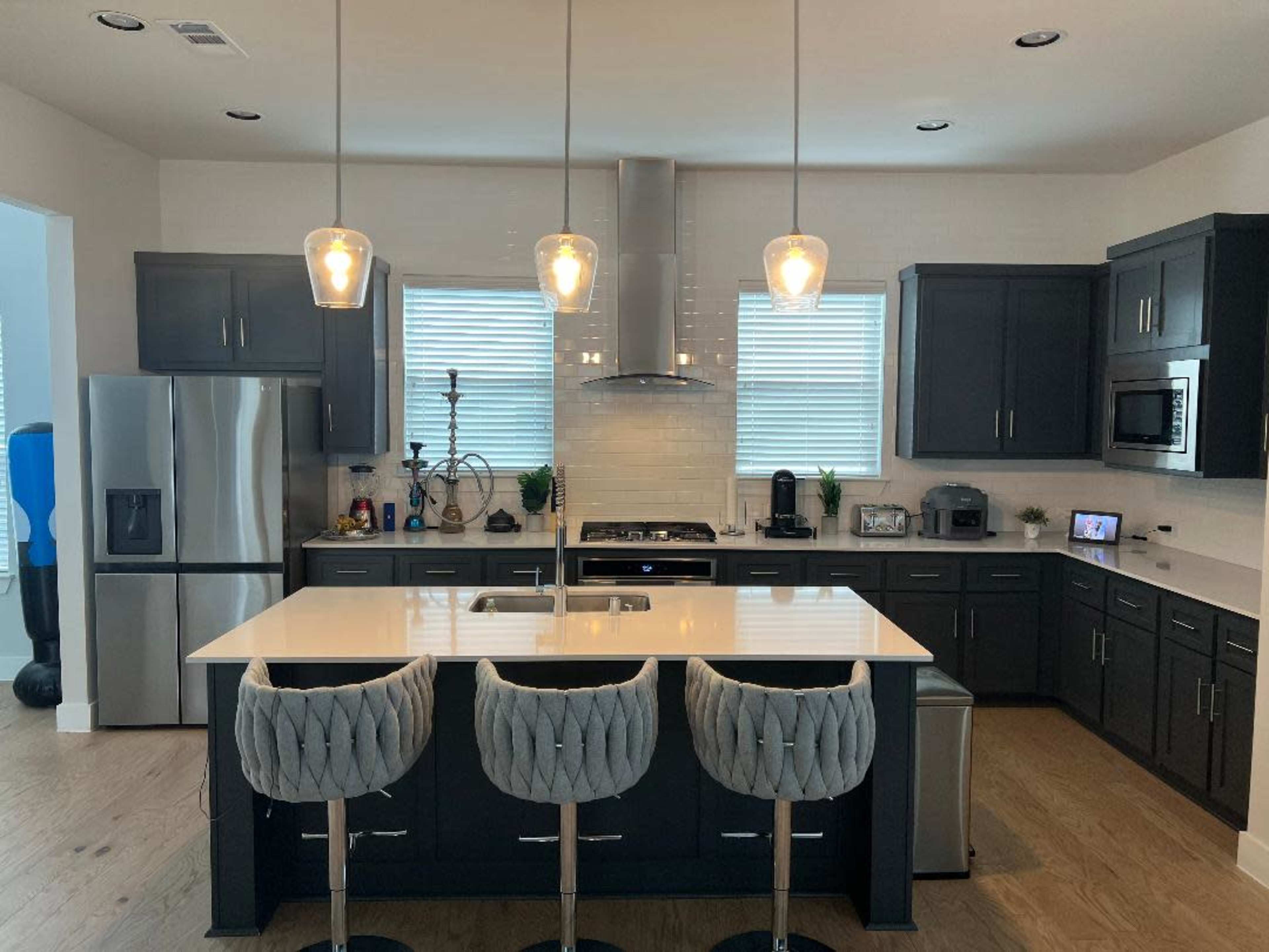 A modern kitchen features a central island with three tufted bar stools, dark cabinetry, stainless steel appliances, and pendant lighting.