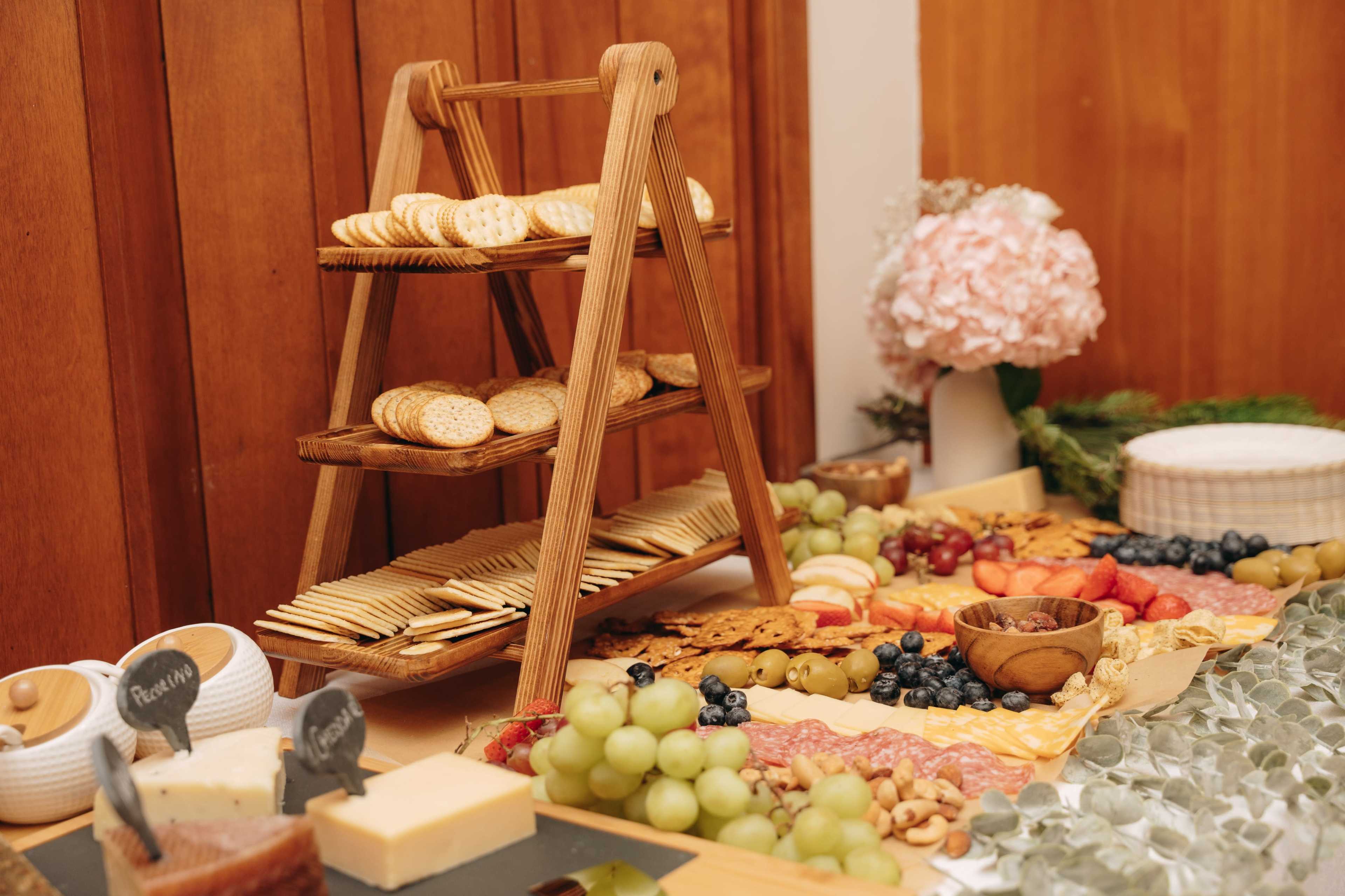 A wooden tiered stand displays an assortment of crackers, while a spread of fruits, cheeses, and nuts is arranged on a table with a floral centerpiece.